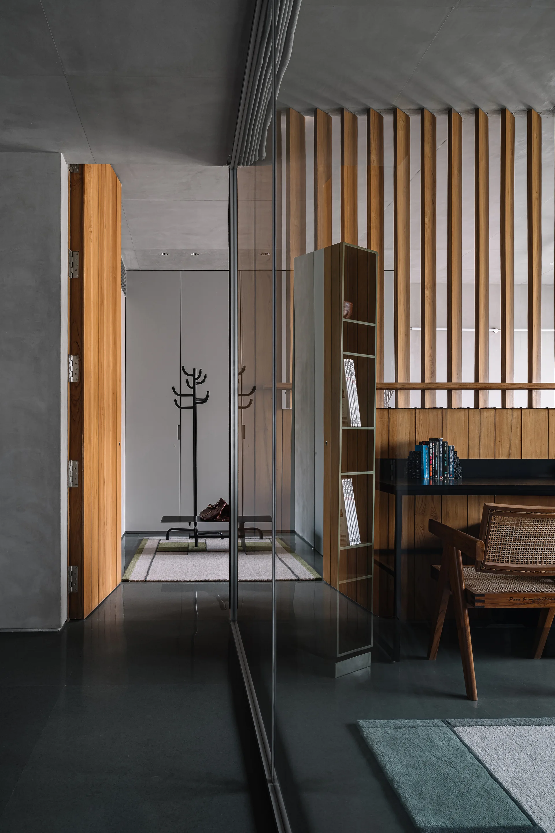 A minimalist home office featuring a glass partition, vertical wood wall slats, and a cane chair set against dark reflective flooring.