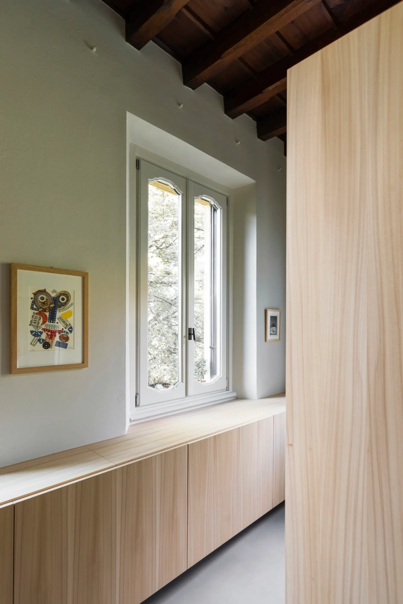 A minimalist room features a long, light-wood built-in cabinet running beneath a white window, offset by dark exposed ceiling beams.