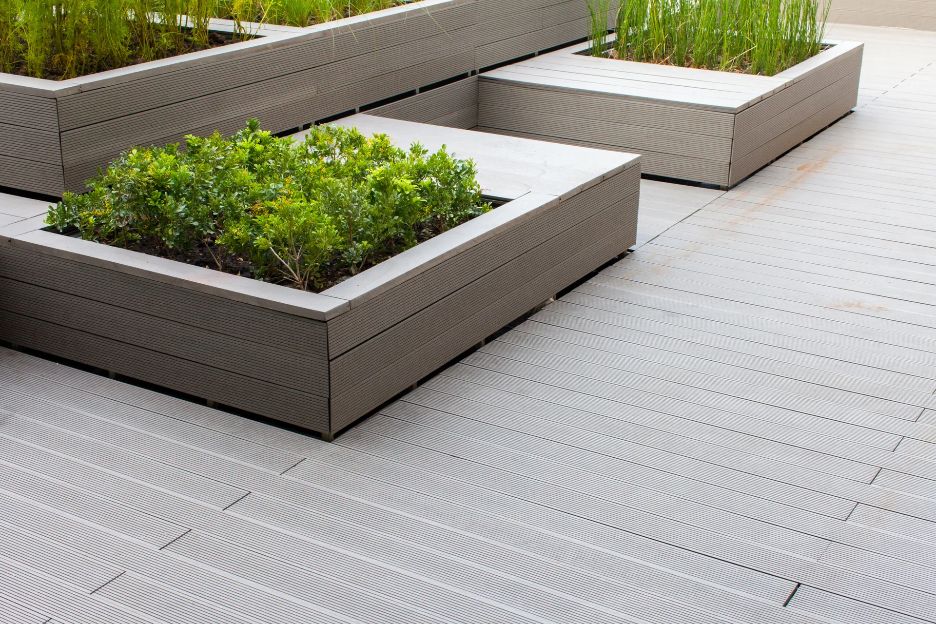 A modern outdoor terrace featuring grey composite wood-toned decking and matching integrated raised planter boxes filled with green leafy plants.