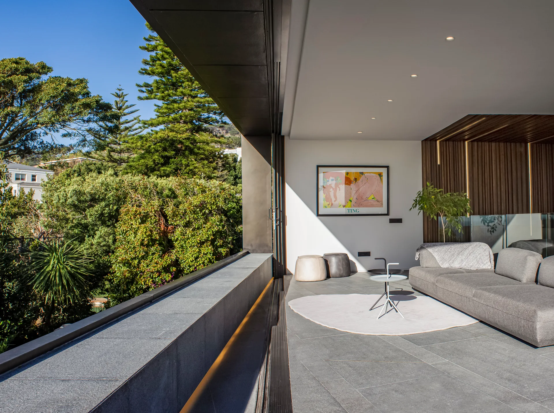 A modern open-air terrace features a gray modular sofa, minimalist side table, and a vertical wood slat wall looking out onto lush green trees.