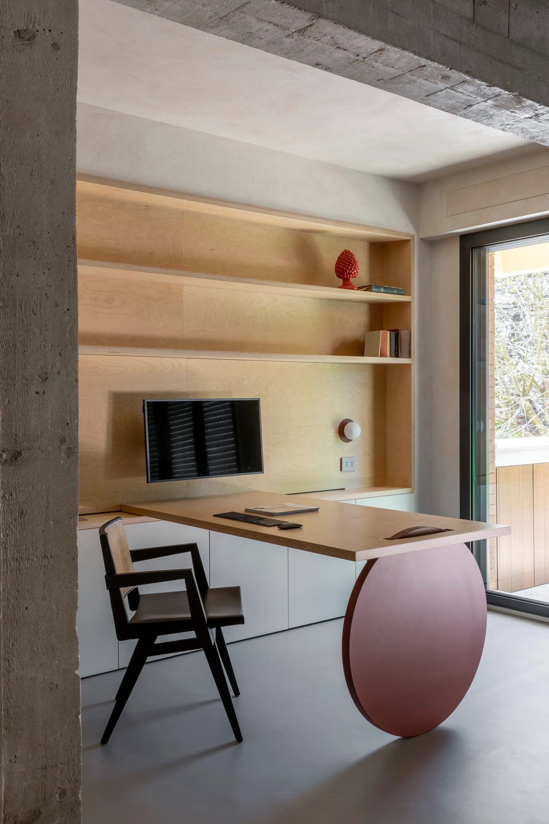 A modern home office with a cantilevered wooden desk supported by a terracotta circular leg, featuring custom wood shelving and a cane-back chair.