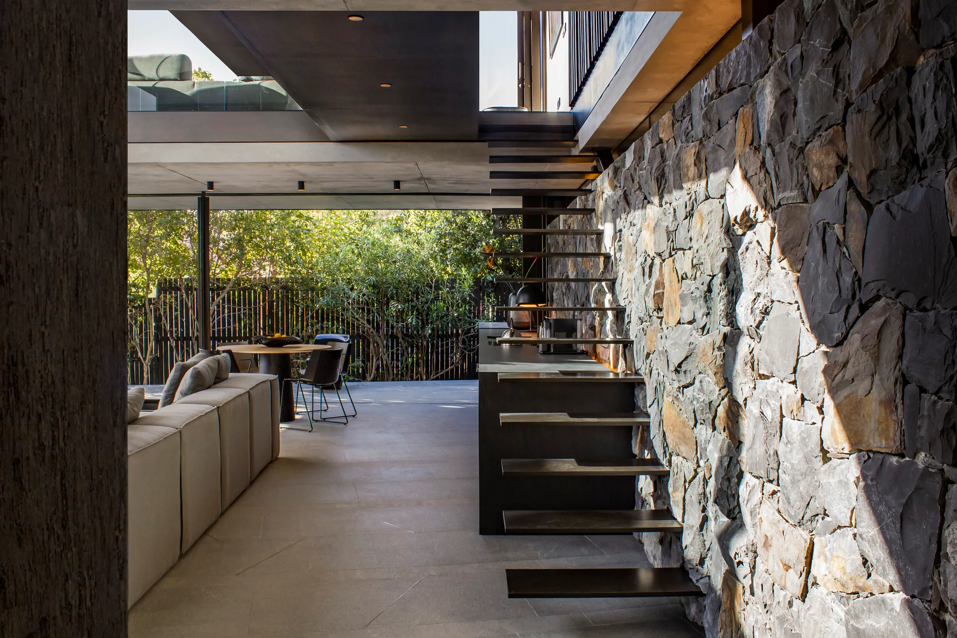 A modern open-plan living room features floating dark metal stairs installed against a rugged grey stone wall.