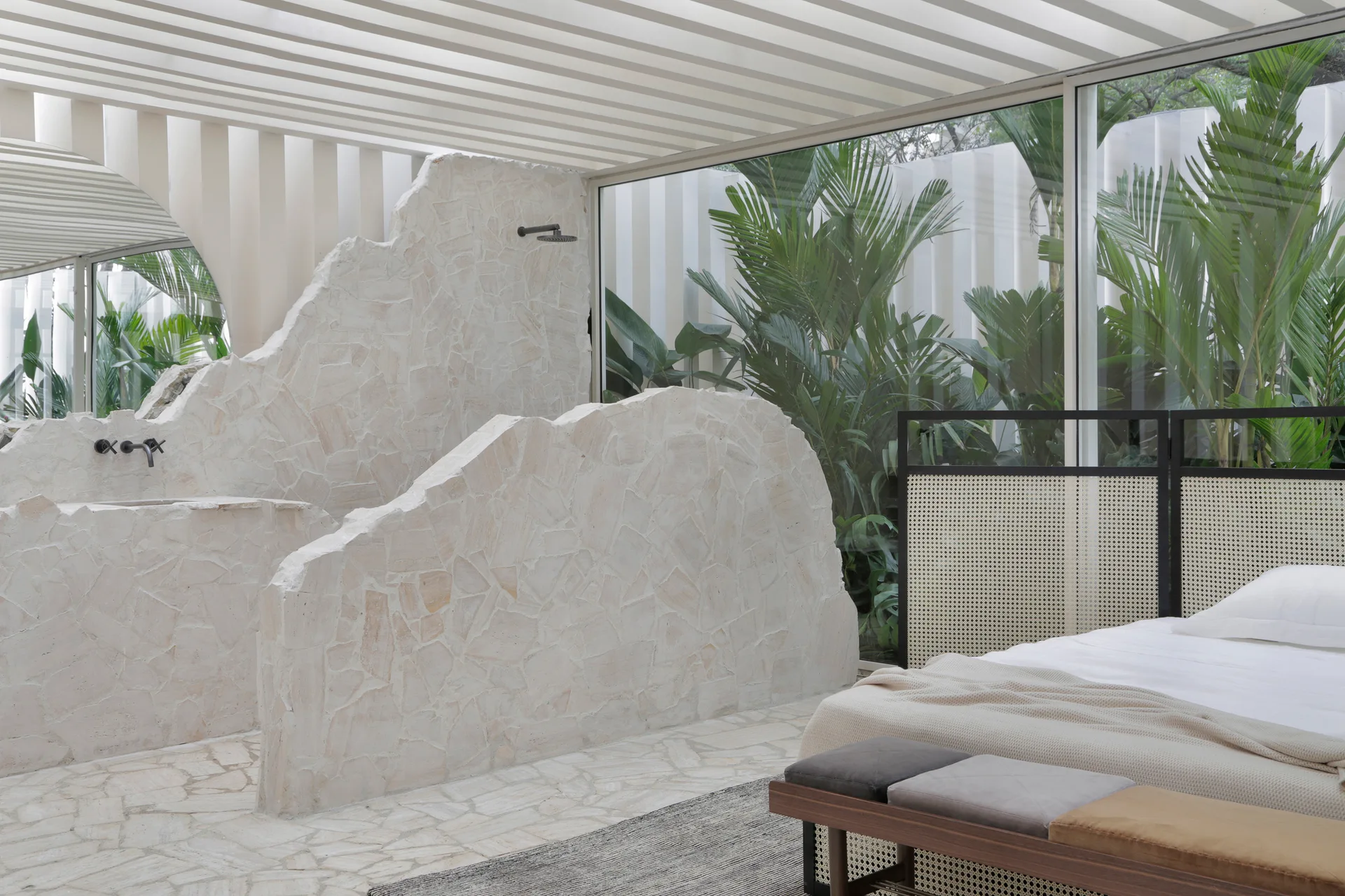 A bedroom features a bed and bench next to a sculptural rough stone partition wall containing an integrated shower, with tropical plants visible outside.