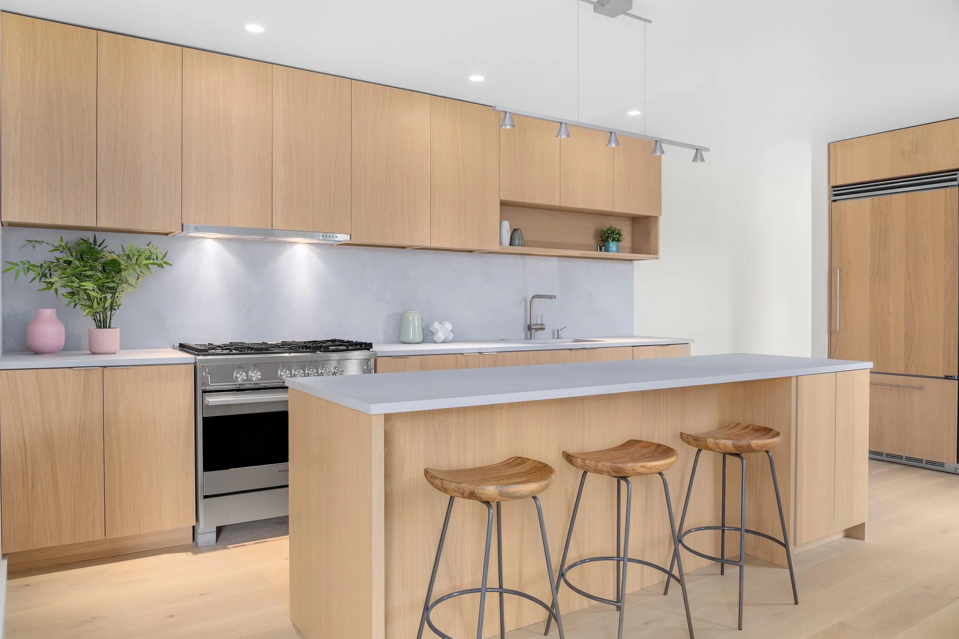 A modern kitchen with light wood flat-panel cabinetry, grey stone-like countertops and backsplash, and three curved wooden bar stools.