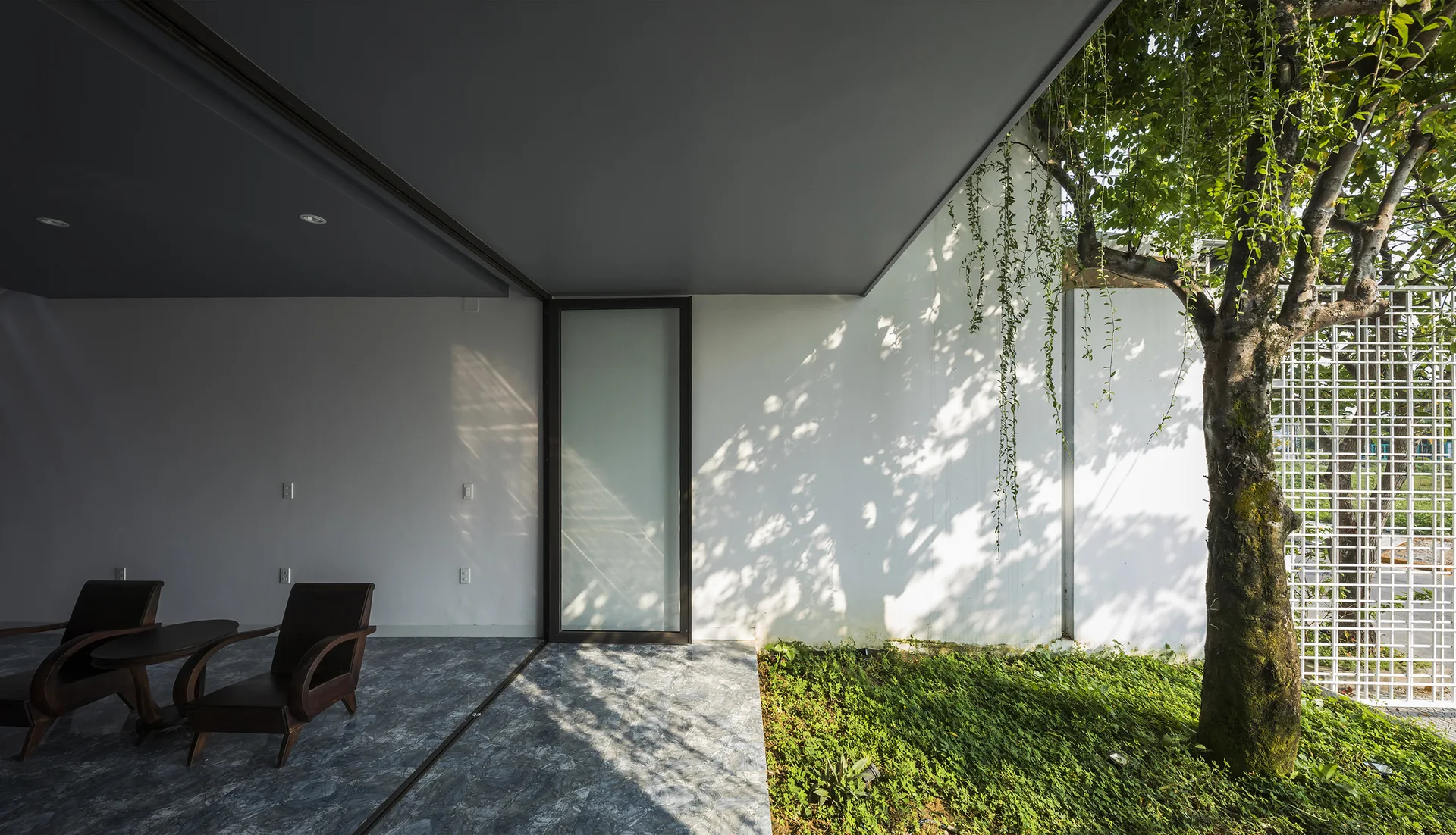 A covered patio with grey stone flooring and dark wood armchairs features a glass sliding door next to a white wall, garden, and tree.