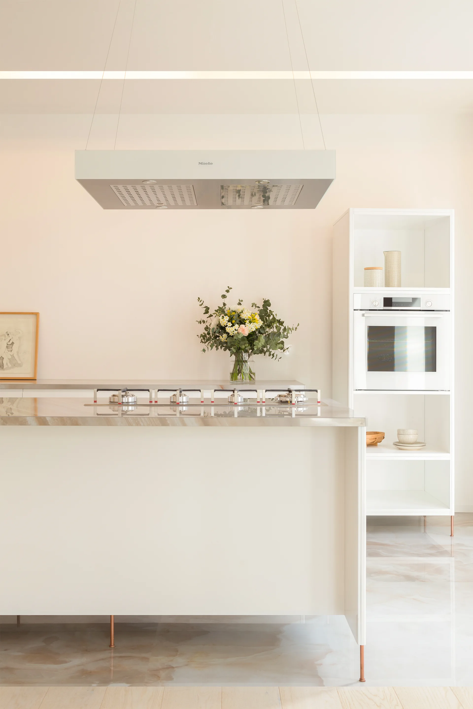 A minimalist white kitchen features a central island with a veined stone countertop, a suspended range hood, and a tall cabinet with an integrated oven.