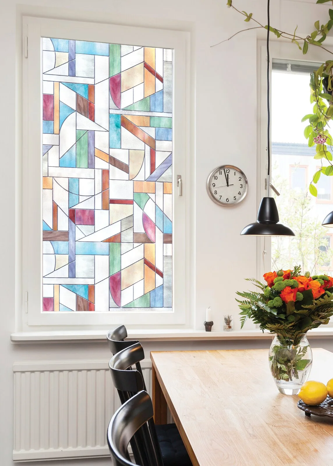 A wooden dining table and black chairs sit near a window covered in a colorful geometric stained glass effect film next to a white wall clock.