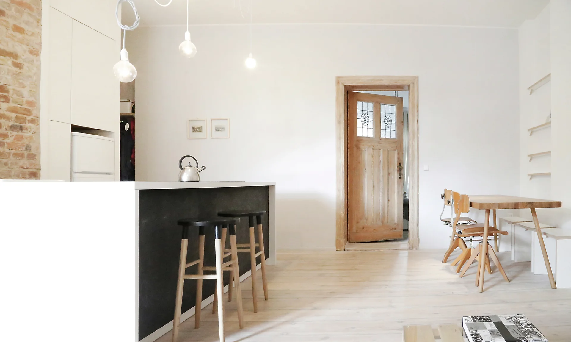 A bright open kitchen and dining space with a black-faced island, natural wood bar stools, light plank flooring, and an old wood door.