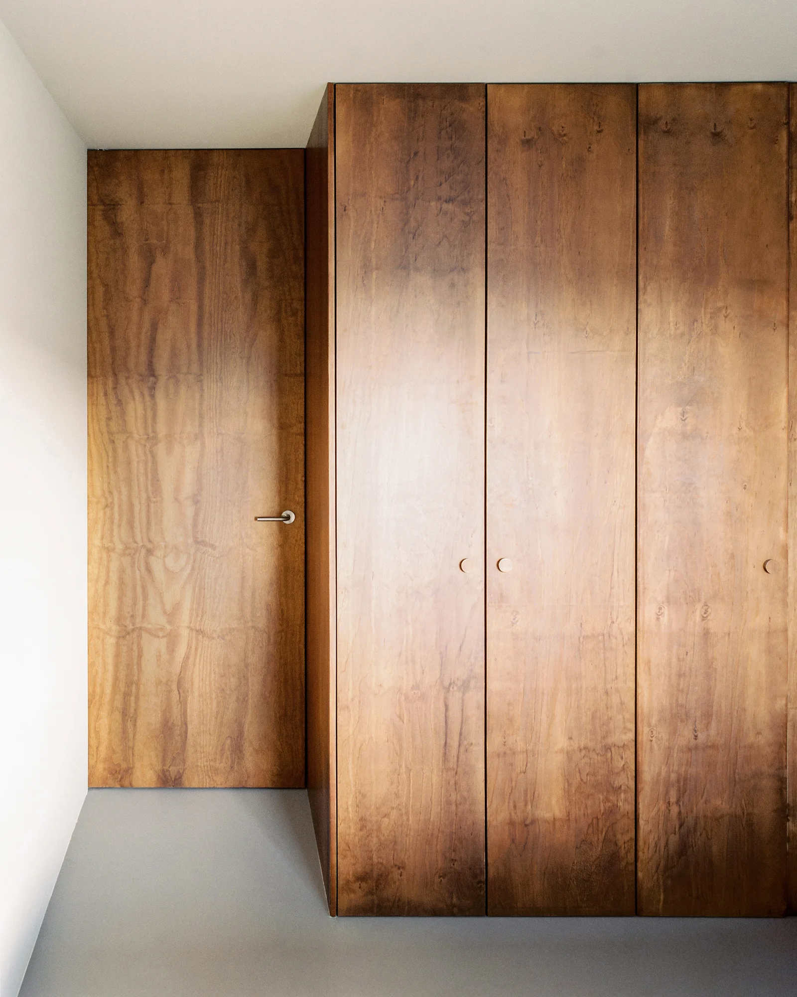 Tall built-in wooden wardrobe cabinets stand next to a matching flush-mounted wood veneer door against a white wall and grey floor.