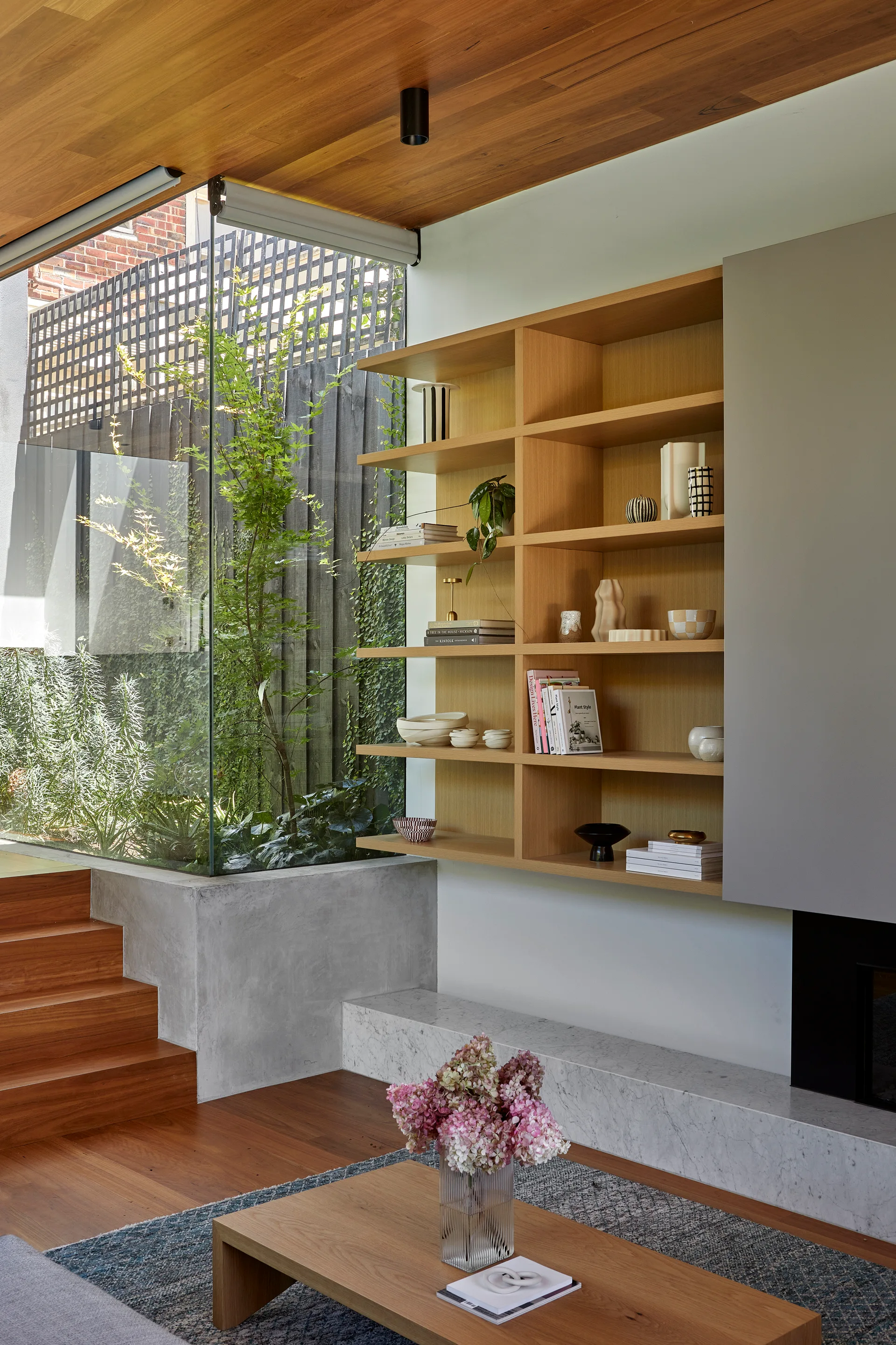 A modern living room features a wood-plank ceiling, built-in oak shelves, a marble hearth fireplace, and a large window looking out onto greenery.
