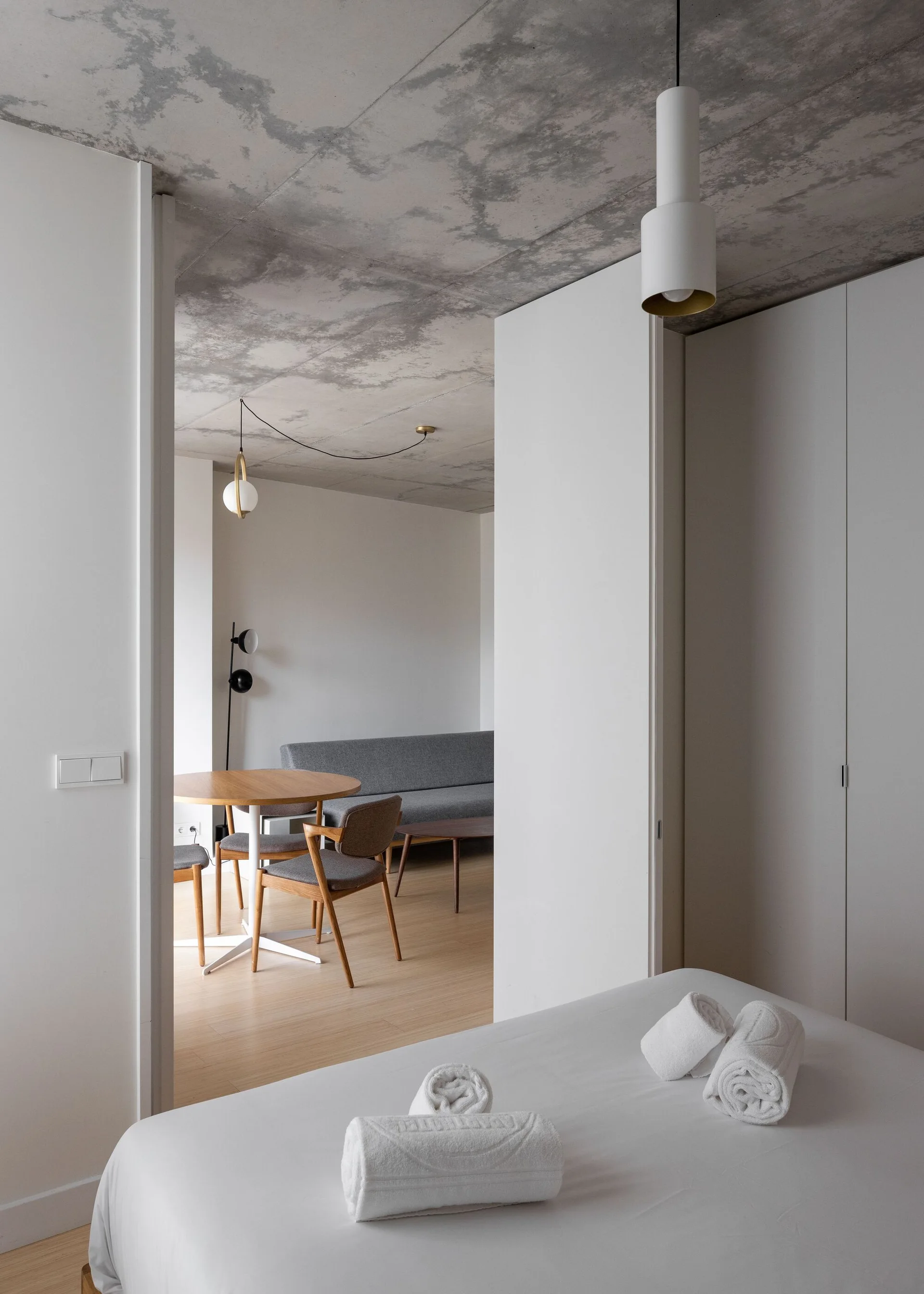 A view from a minimalist bedroom looking into a living space with a round wood dining table, grey sofa, and raw concrete ceiling.