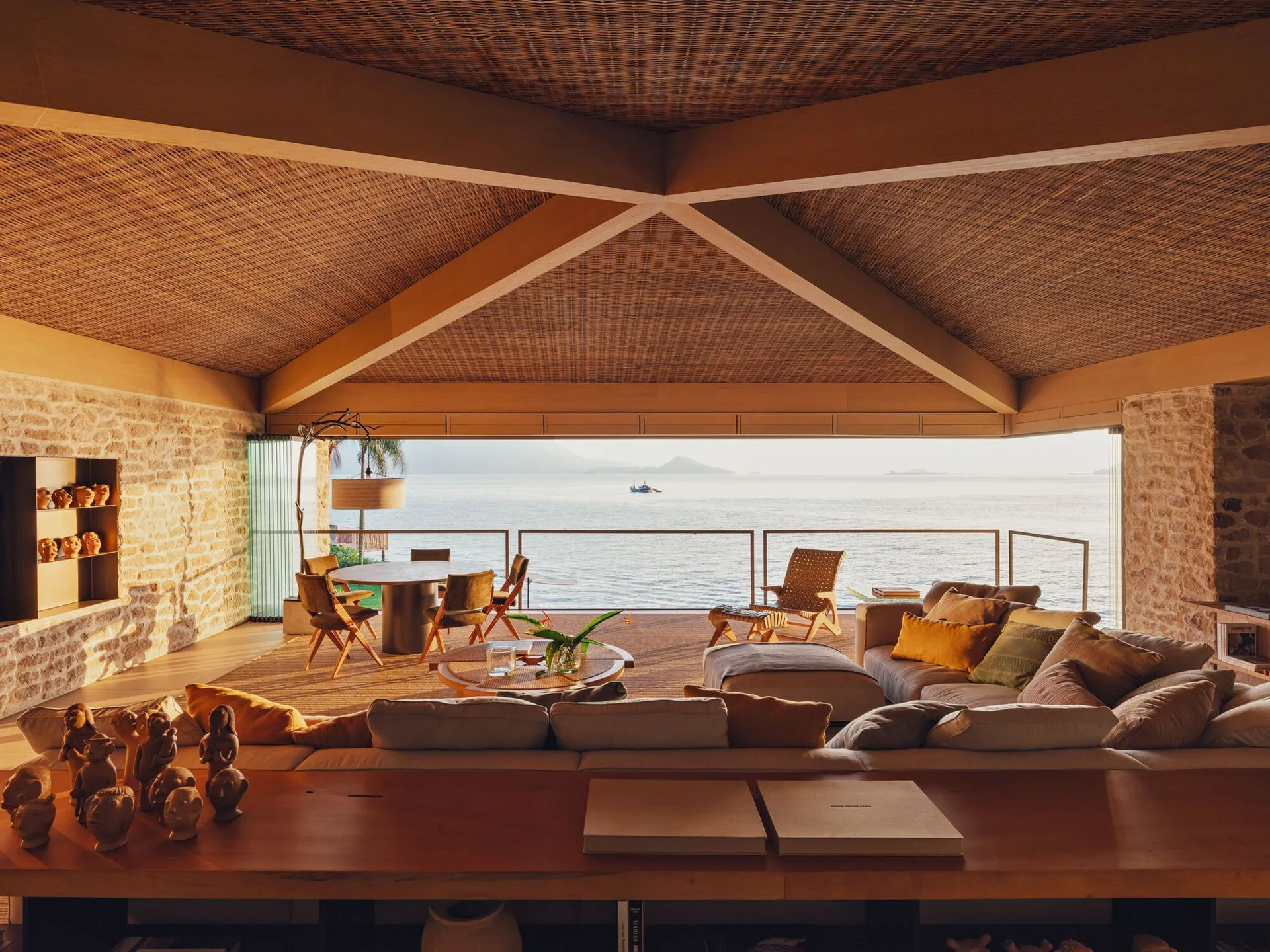 An open-concept living room features a patterned wooden vaulted ceiling, a modular sofa, stone walls, and a wide balcony overlooking the ocean.