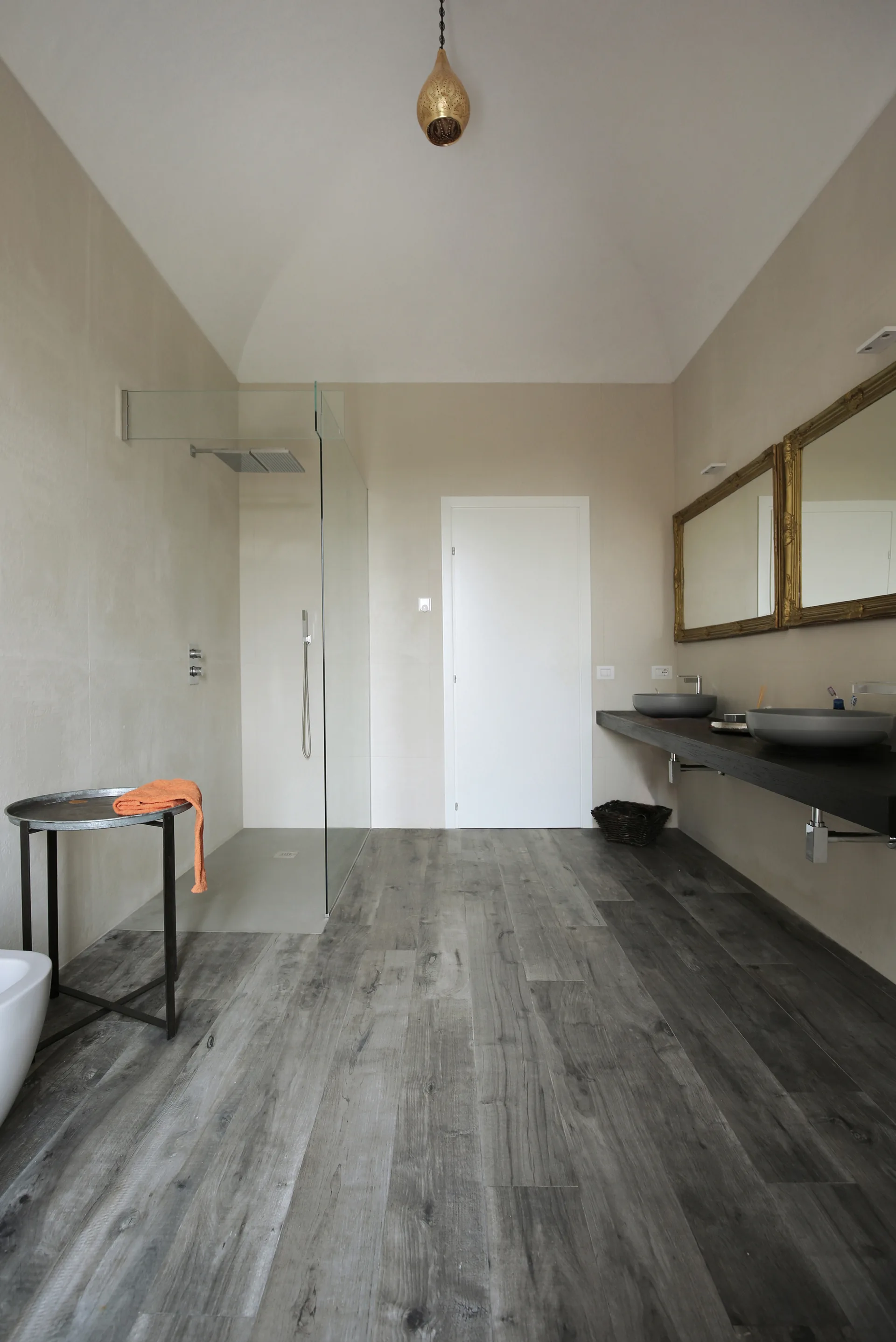 A bathroom featuring a long dark wood floating vanity with double vessel sinks, gold-framed mirrors, and a glass walk-in shower.