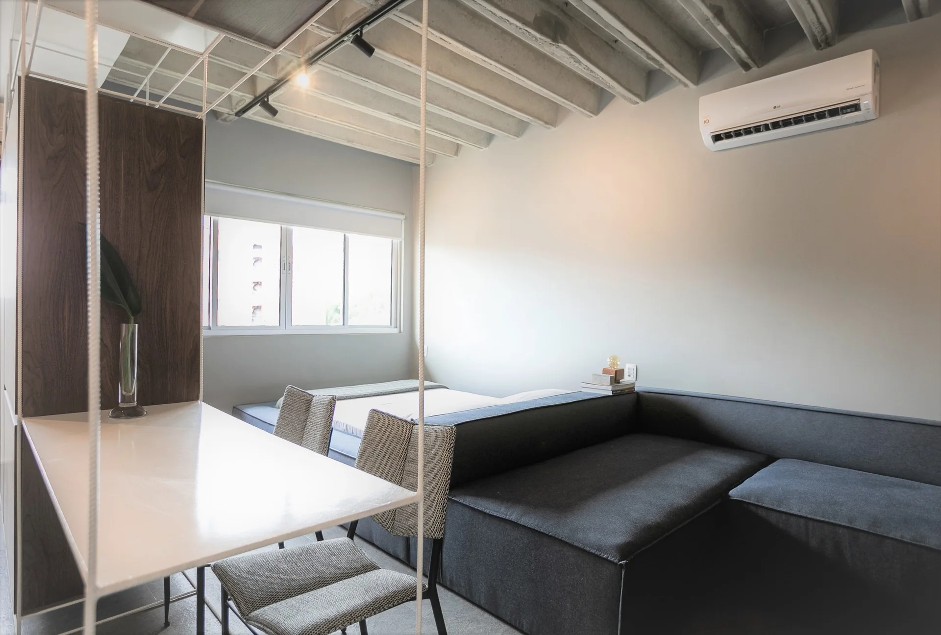 A modern studio apartment features a suspended white table, gray dining chairs, a dark sectional sofa, and exposed concrete ceiling beams.