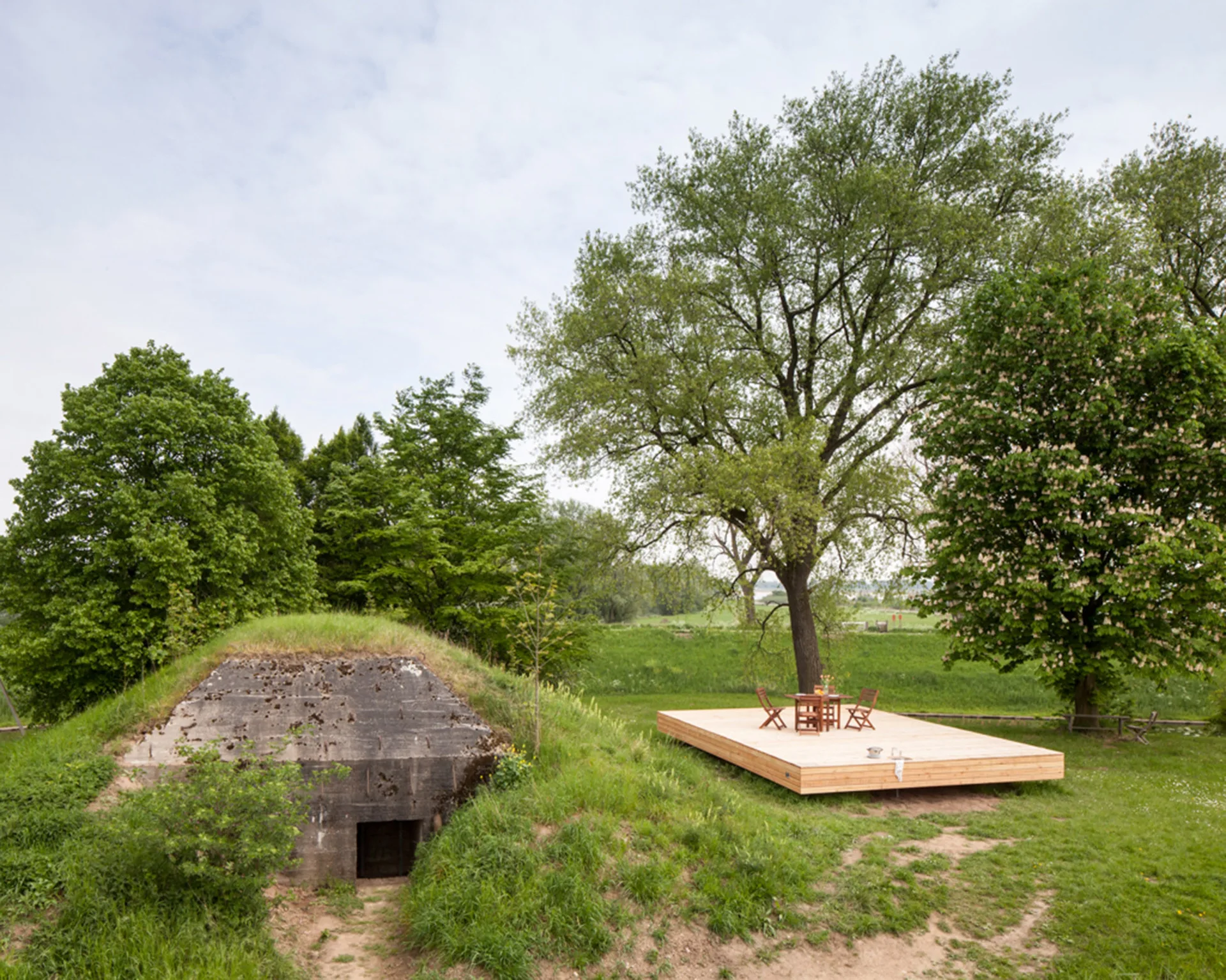 A raised rectangular light wood platform with a dining table and chairs sits in a grassy field next to a grass-covered concrete bunker.