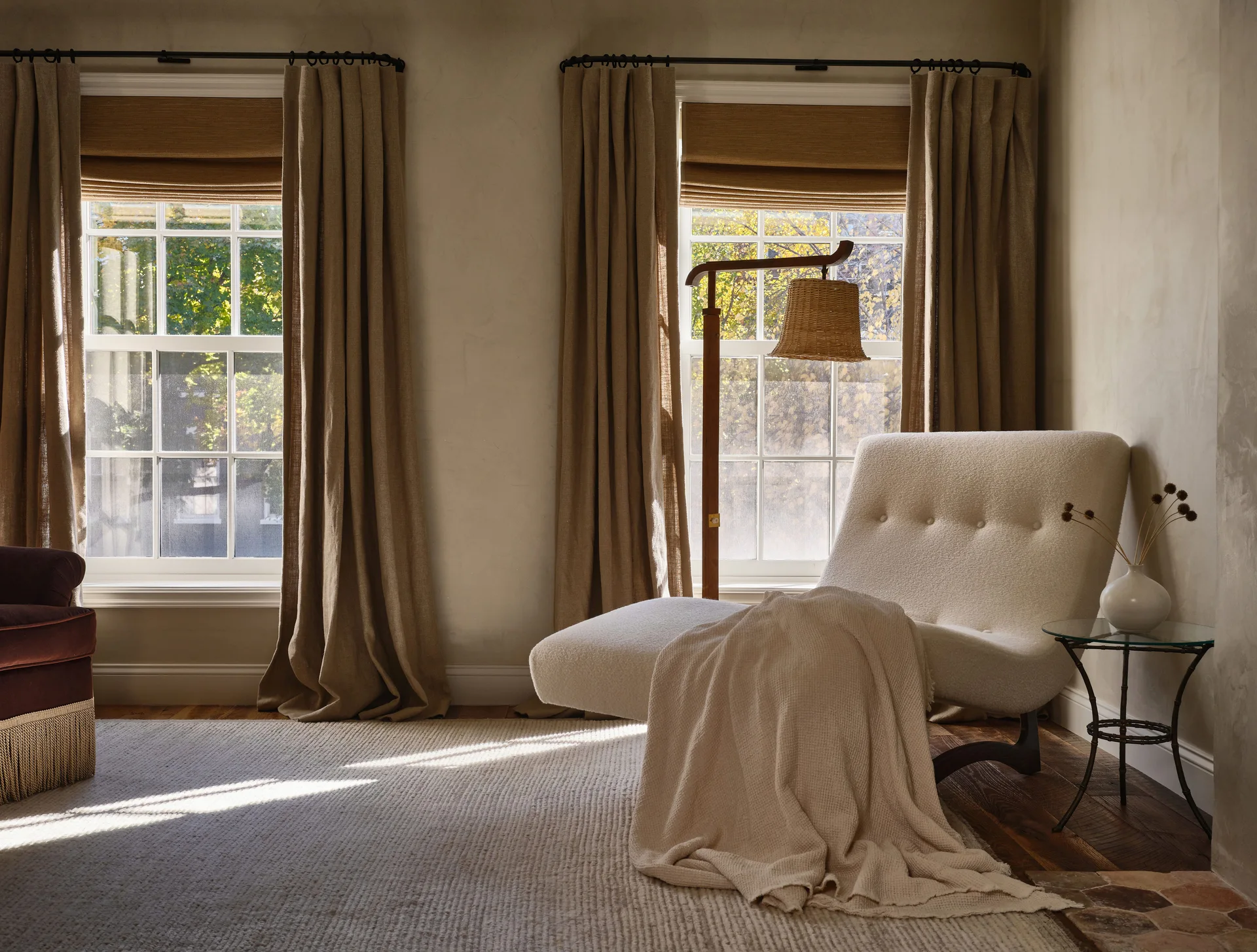 A cream chaise lounge with a throw blanket sits on a textured rug near two windows featuring beige linen curtains and woven shades.