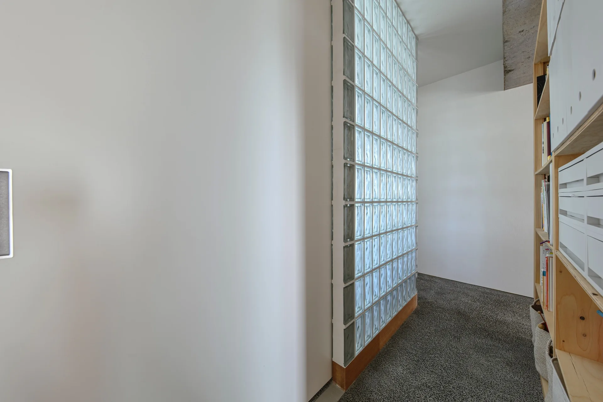 A modern white hallway features a tall translucent glass block partition wall next to light wood open shelving and grey speckled flooring.