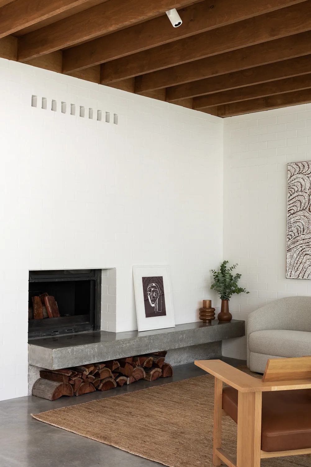 A minimalist living room featuring a white painted brick wall, a low concrete fireplace hearth, and exposed wooden ceiling beams.