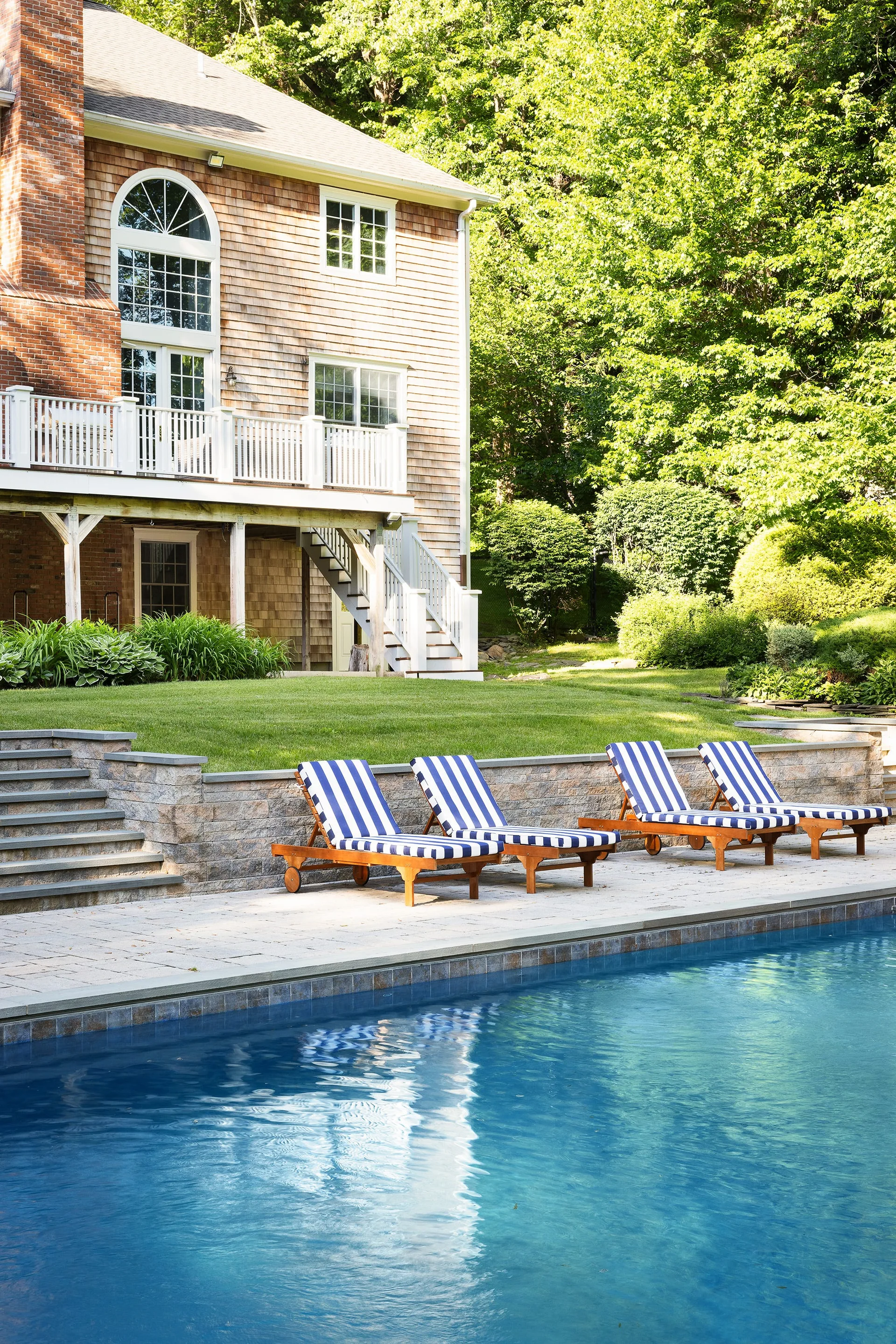 Four wooden lounge chairs with blue and white striped cushions sit on a stone patio next to an in-ground swimming pool with a house in the background.