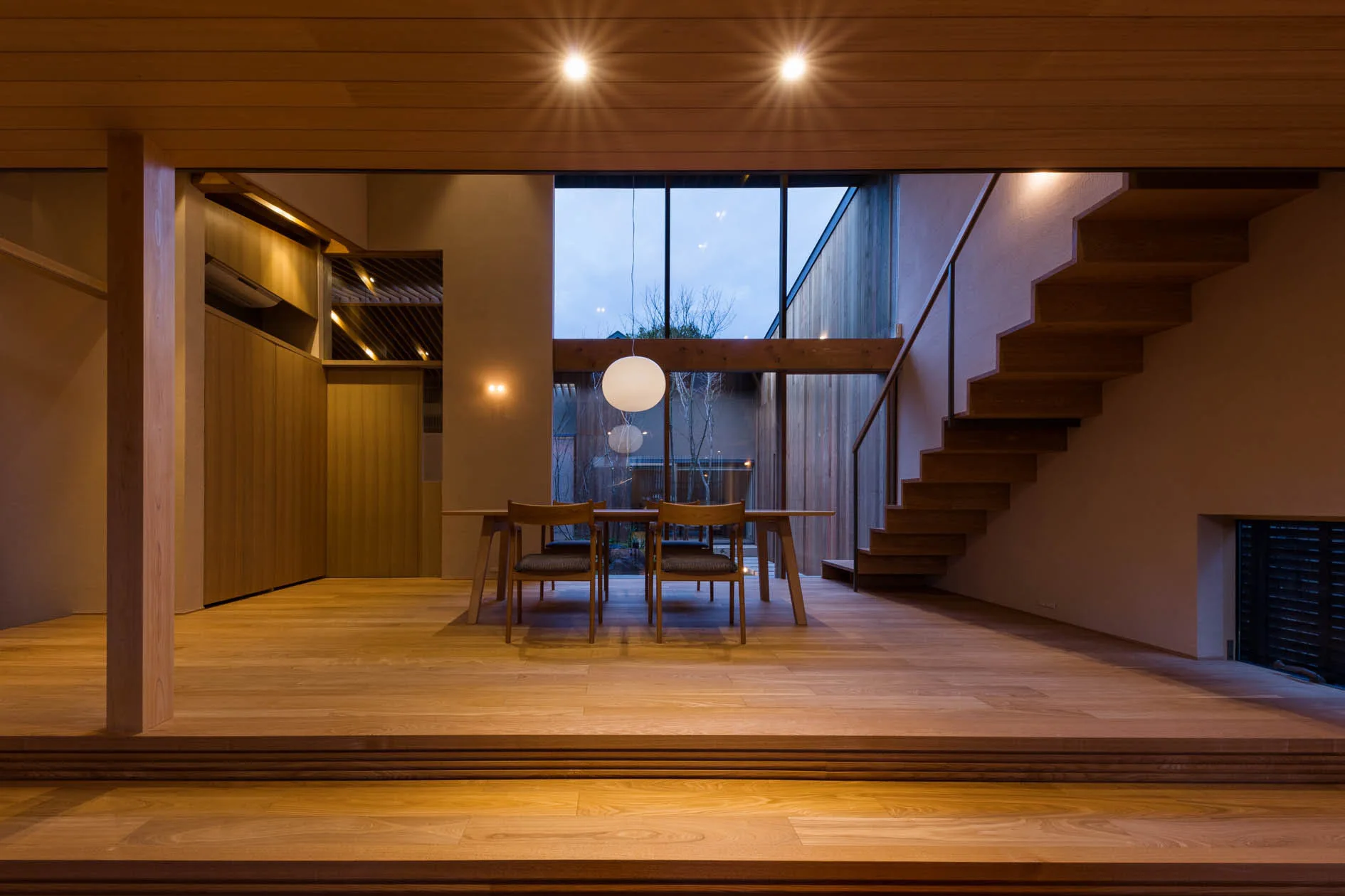 A minimalist dining room features a wooden table and chairs, an open floating staircase, and large glass windows overlooking an outdoor courtyard.