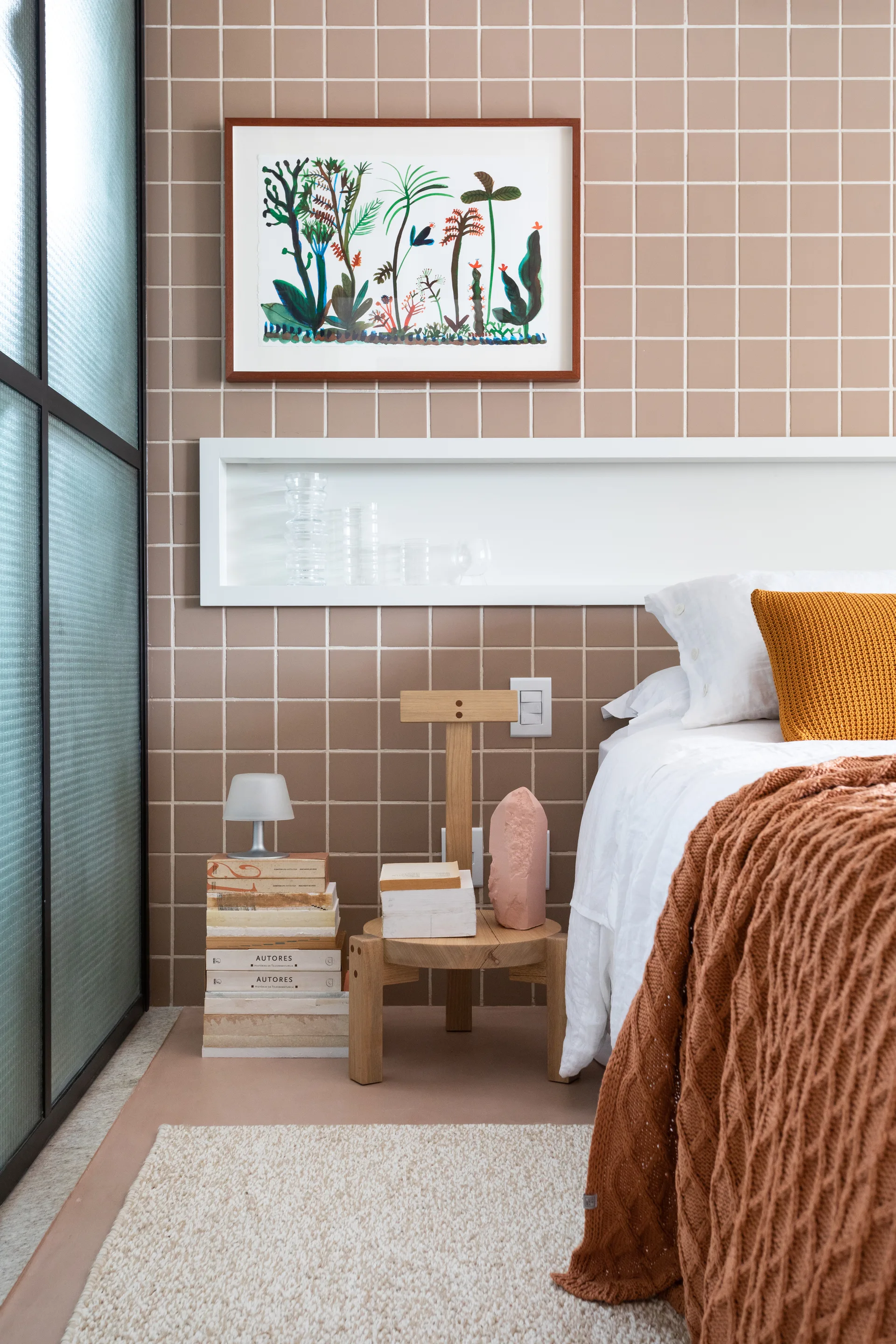 A bedroom features a brown square-tiled wall with a recessed white niche shelf above a wooden side table, stack of books, and white bed.