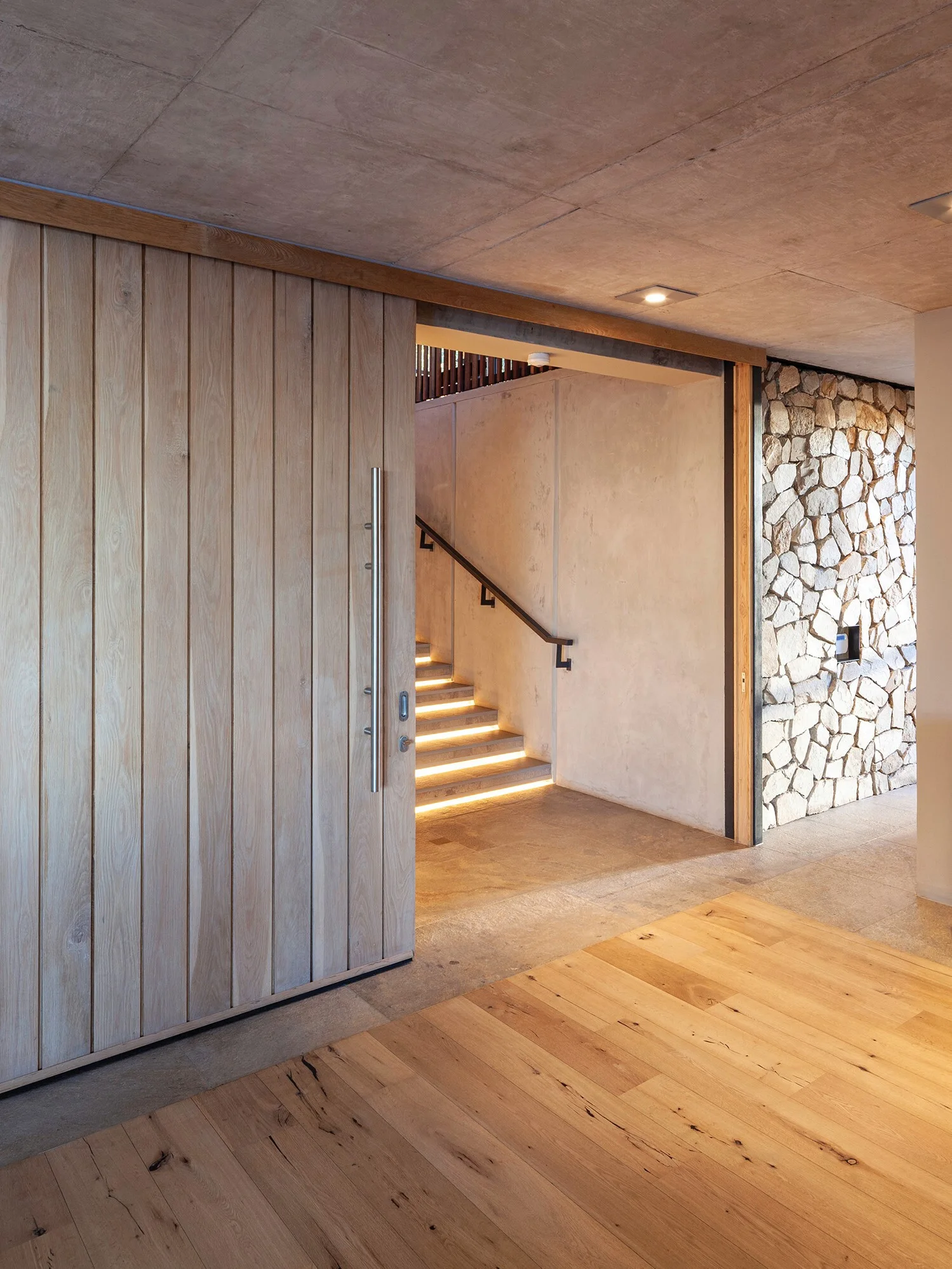 A modern interior entryway featuring a large wood sliding door, a concrete ceiling, and a staircase with integrated under-tread lighting.