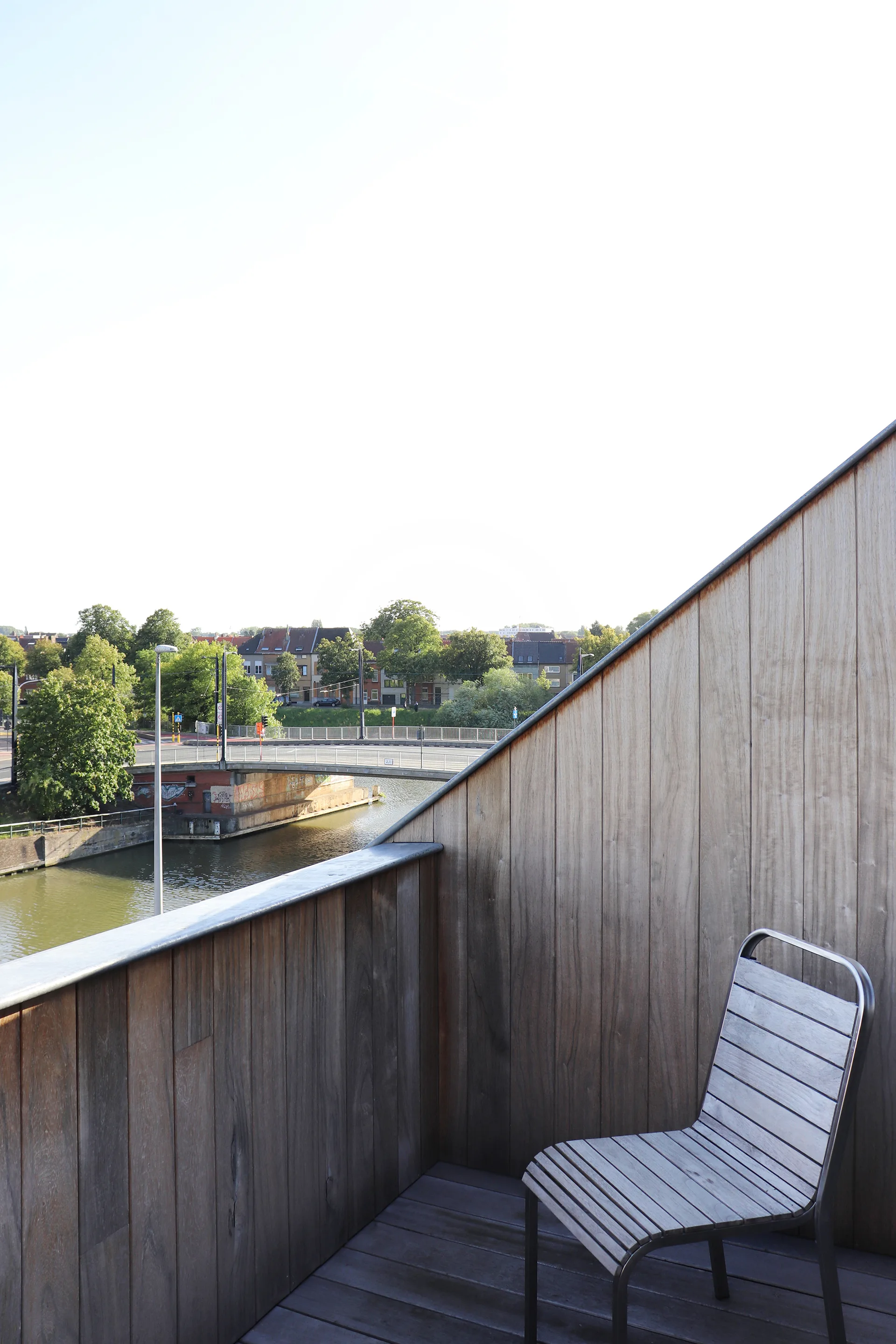 A minimalist wooden balcony features vertical timber cladding, a slatted chair, and a view of a river and bridge in the distance.