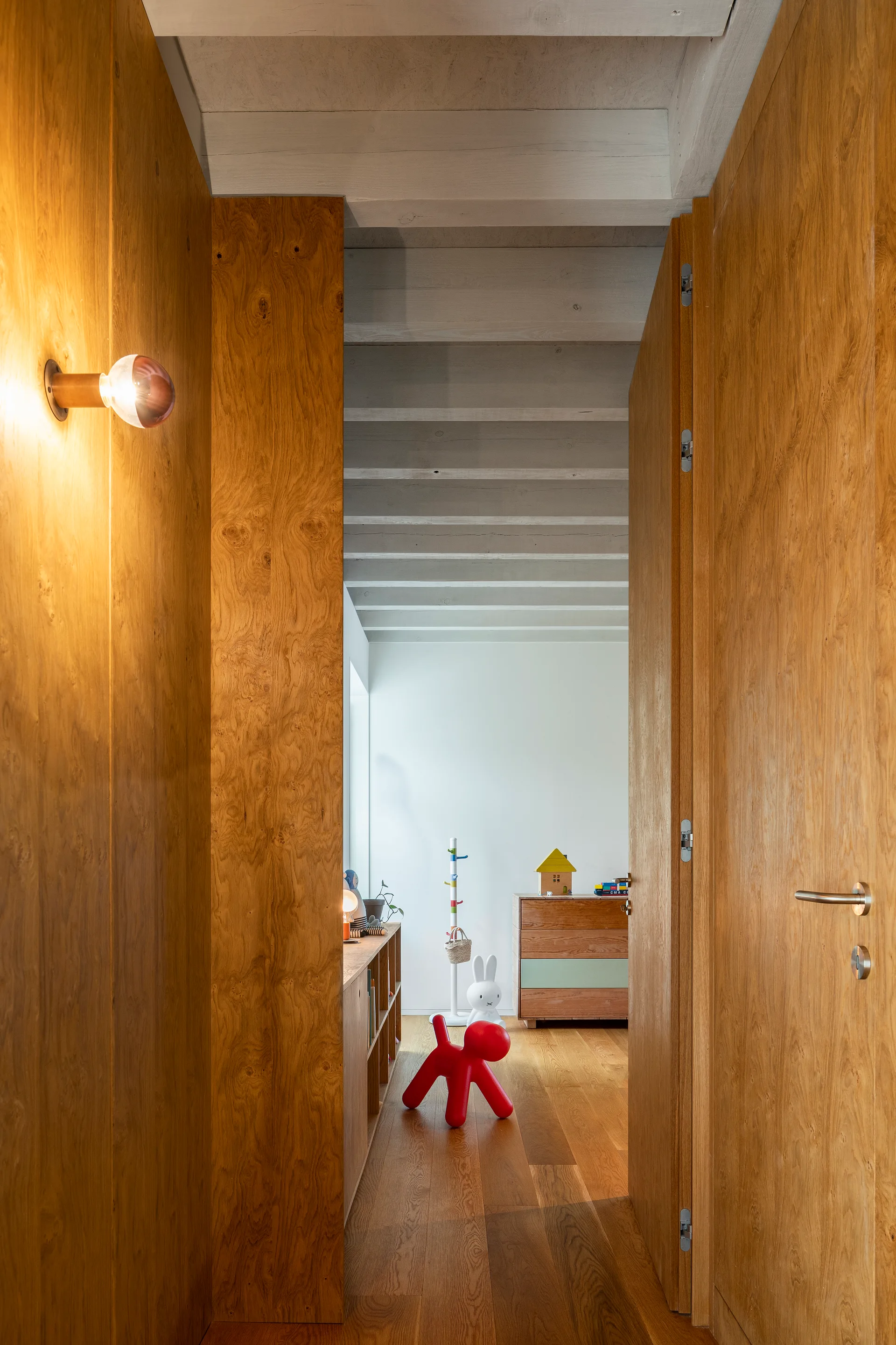 A view through a warm wood paneled doorway into a bright children's room featuring white walls, exposed ceiling beams, and a red toy dog.