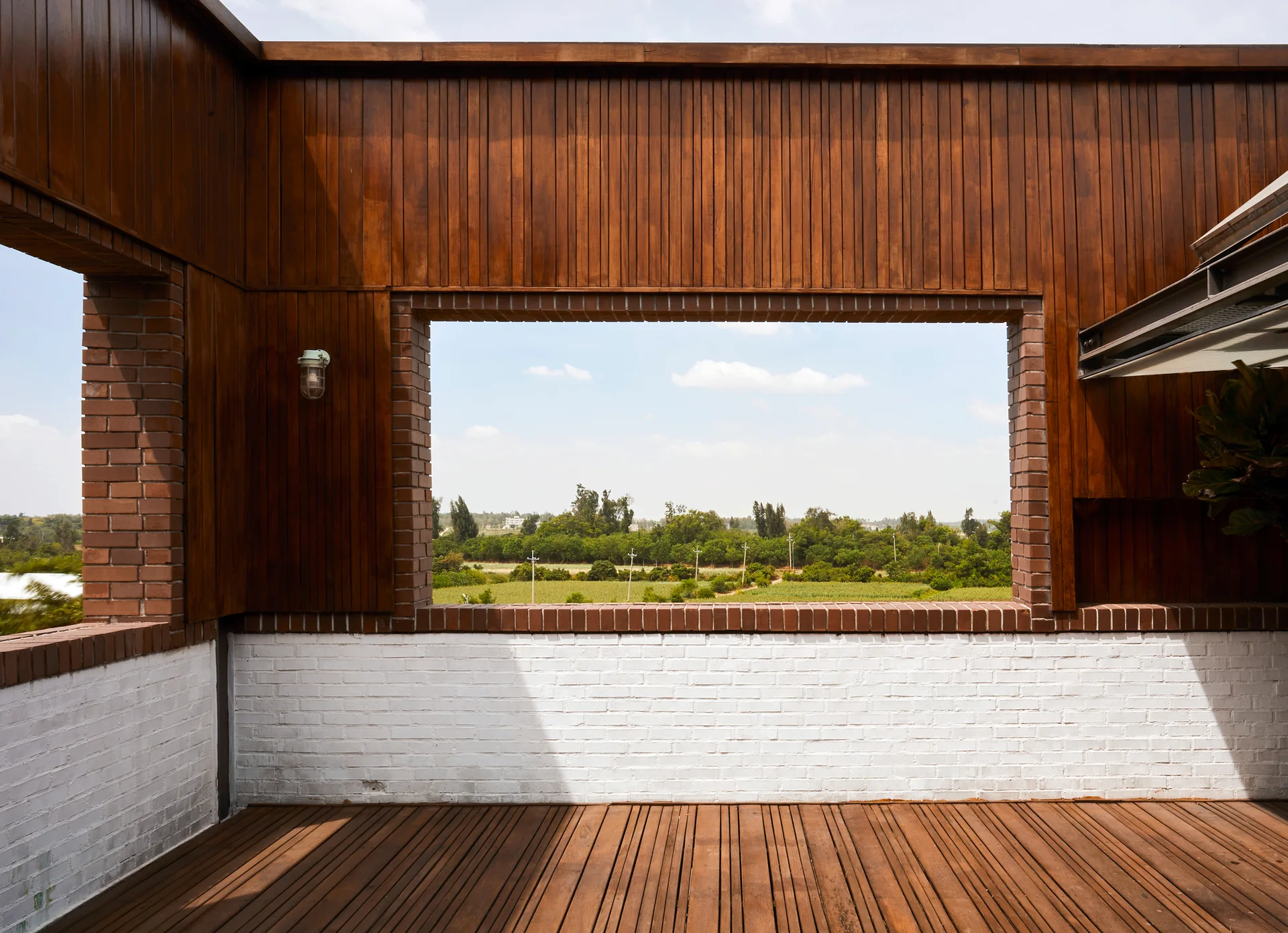 An outdoor wooden terrace deck features vertical wood paneling, a white painted brick wall, and a large open rectangular window framing a landscape view.