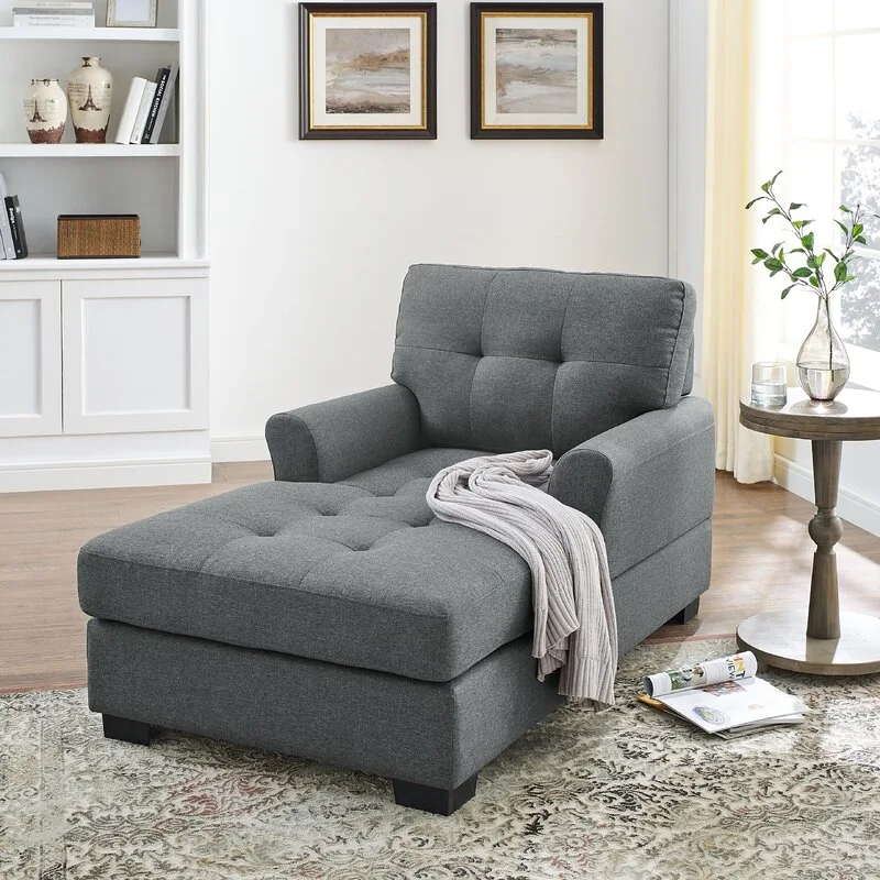 A grey tufted fabric chaise lounge sits on an ornate patterned rug next to a wooden side table and white built-in shelving.