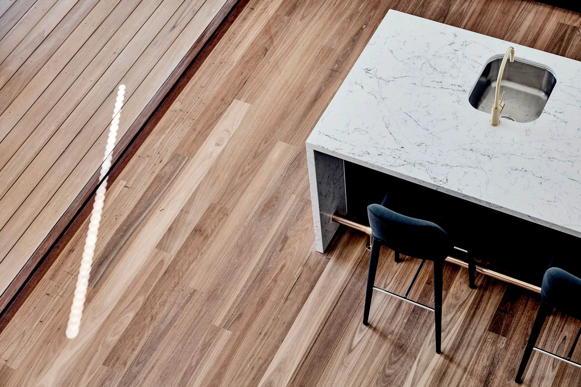 A high-angle view of a white marble kitchen island with a brass faucet, stainless steel sink, and dark stools on warm wood plank floors.