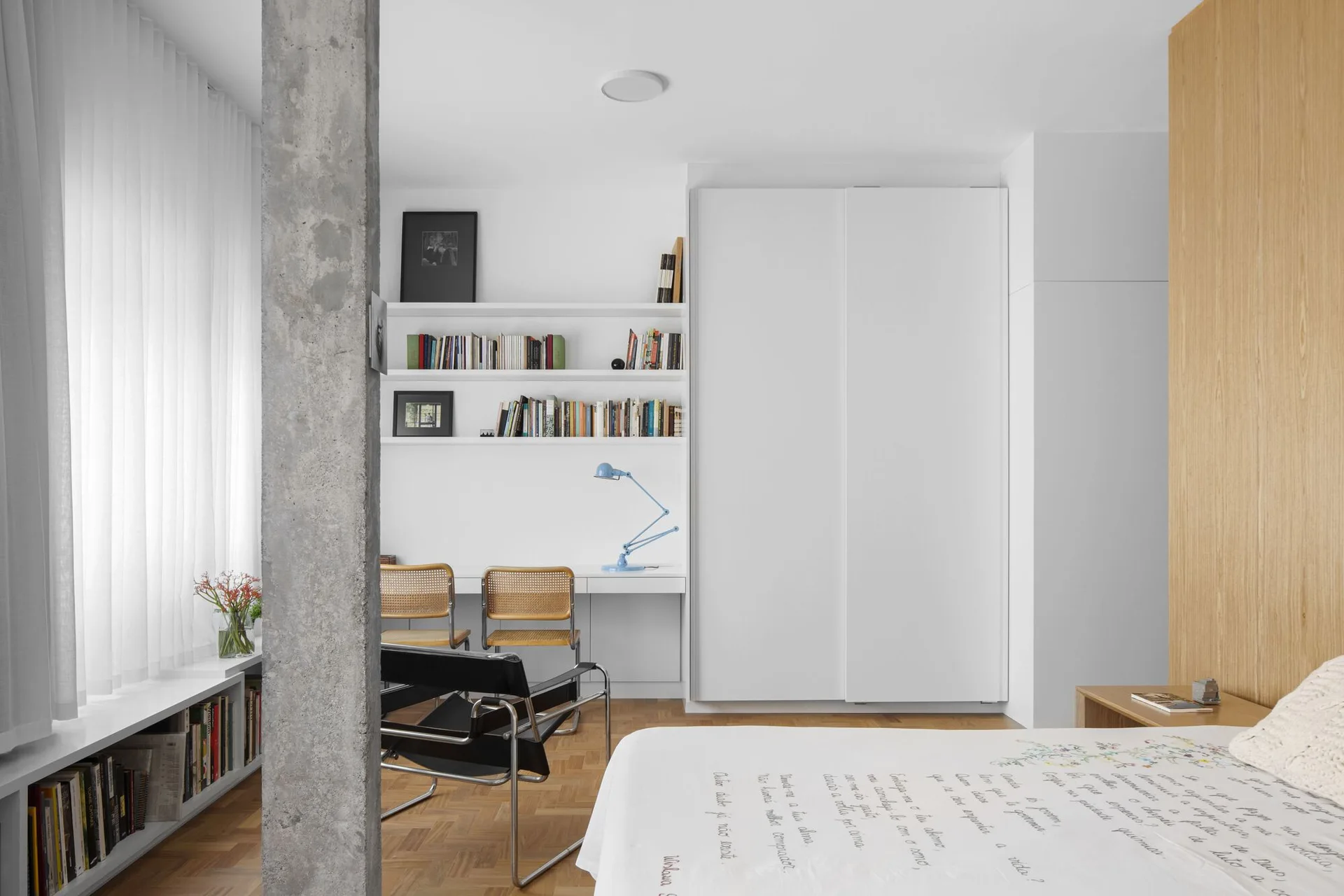 A modern bedroom features a raw concrete column, a built-in white desk with shelving, and a bed with text-patterned bedding.