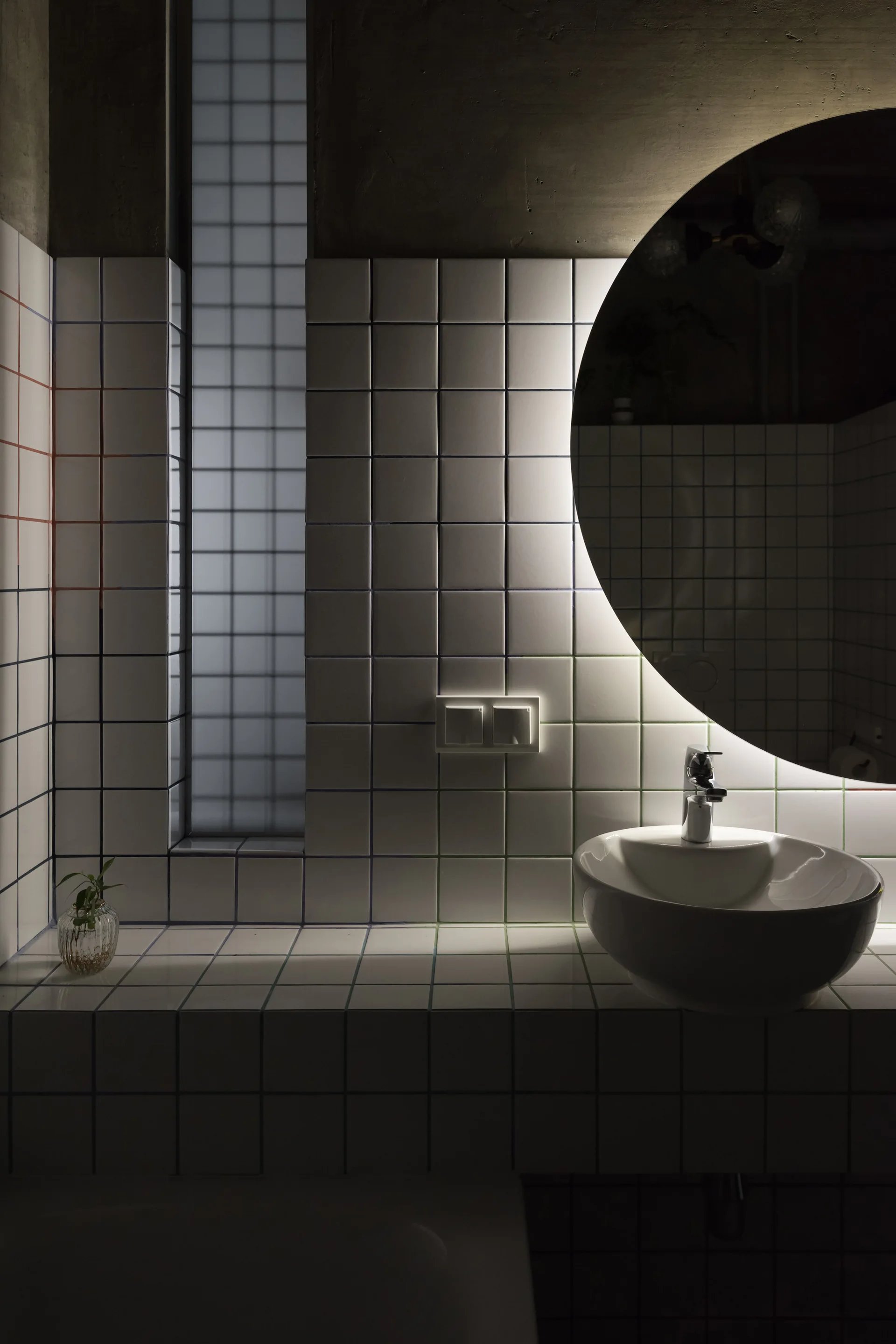 A modern bathroom vanity features white square tiles, a round backlit mirror, a white ceramic vessel sink, and a narrow frosted window.