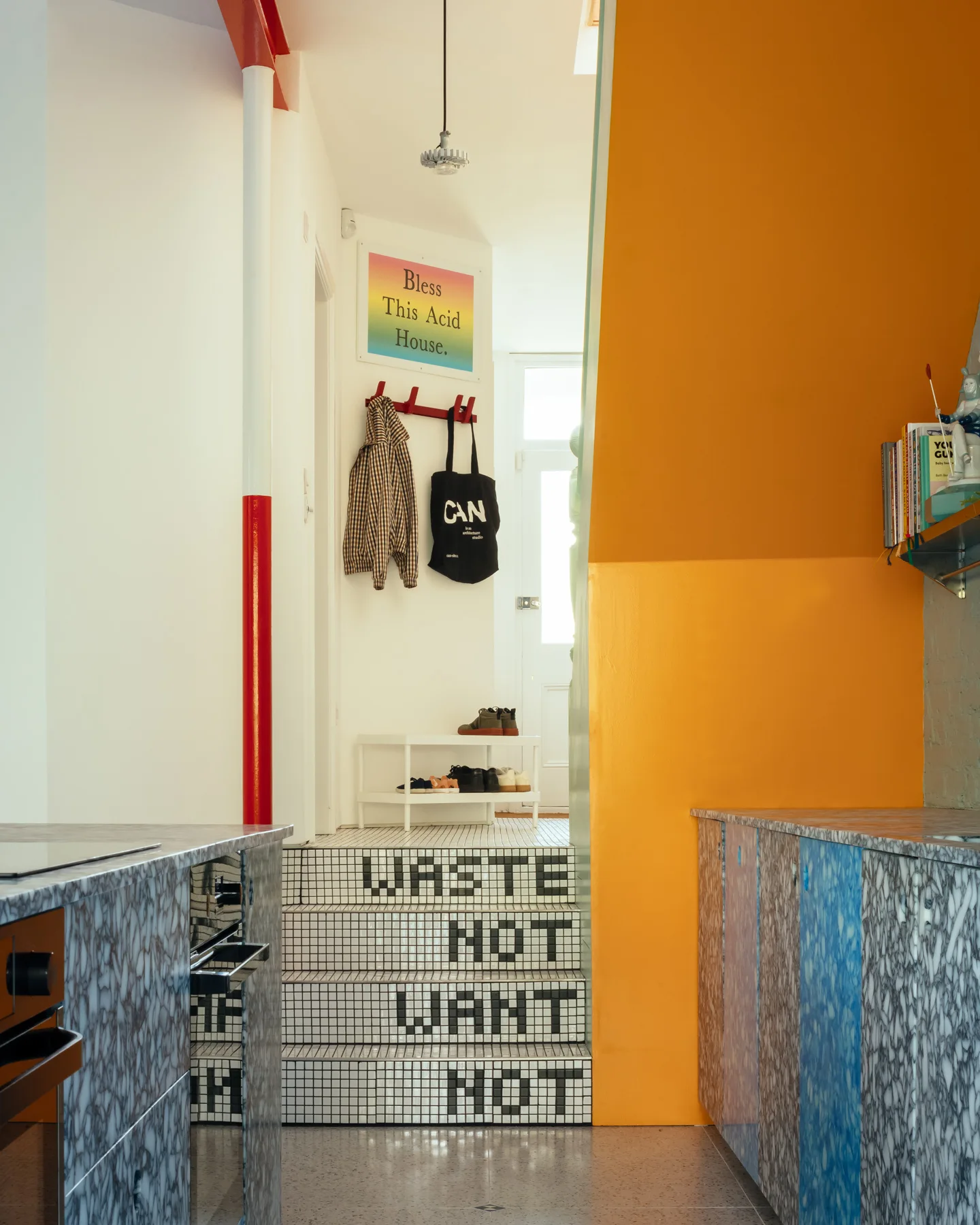 An eclectic entryway features white mosaic tiled stairs spelled out with graphic black text, flanked by marble-patterned cabinets and a bright orange wall.