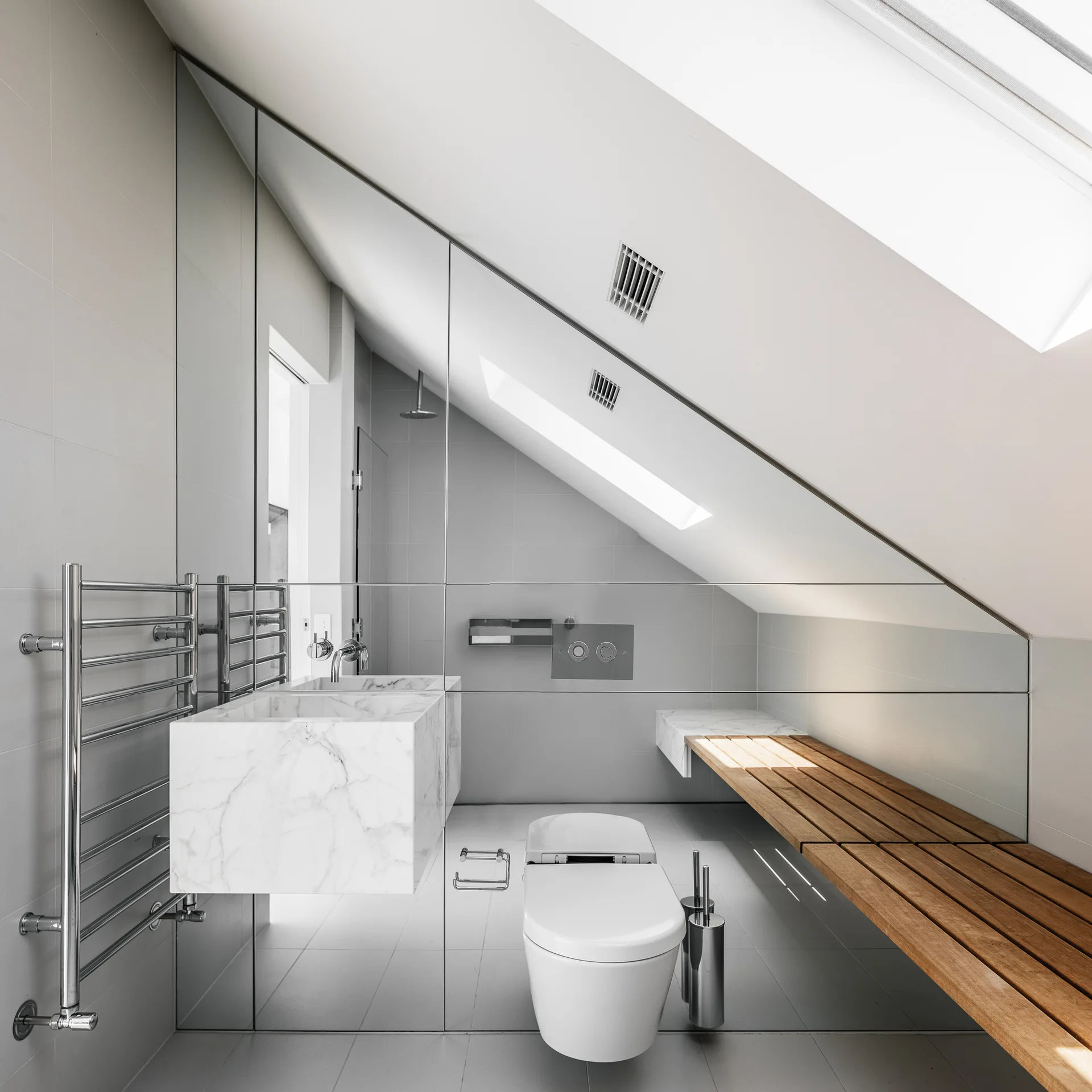 An attic bathroom features a wall-mounted marble sink, a white toilet, a slatted wooden bench, and a wall-to-wall mirror under a sloped ceiling.