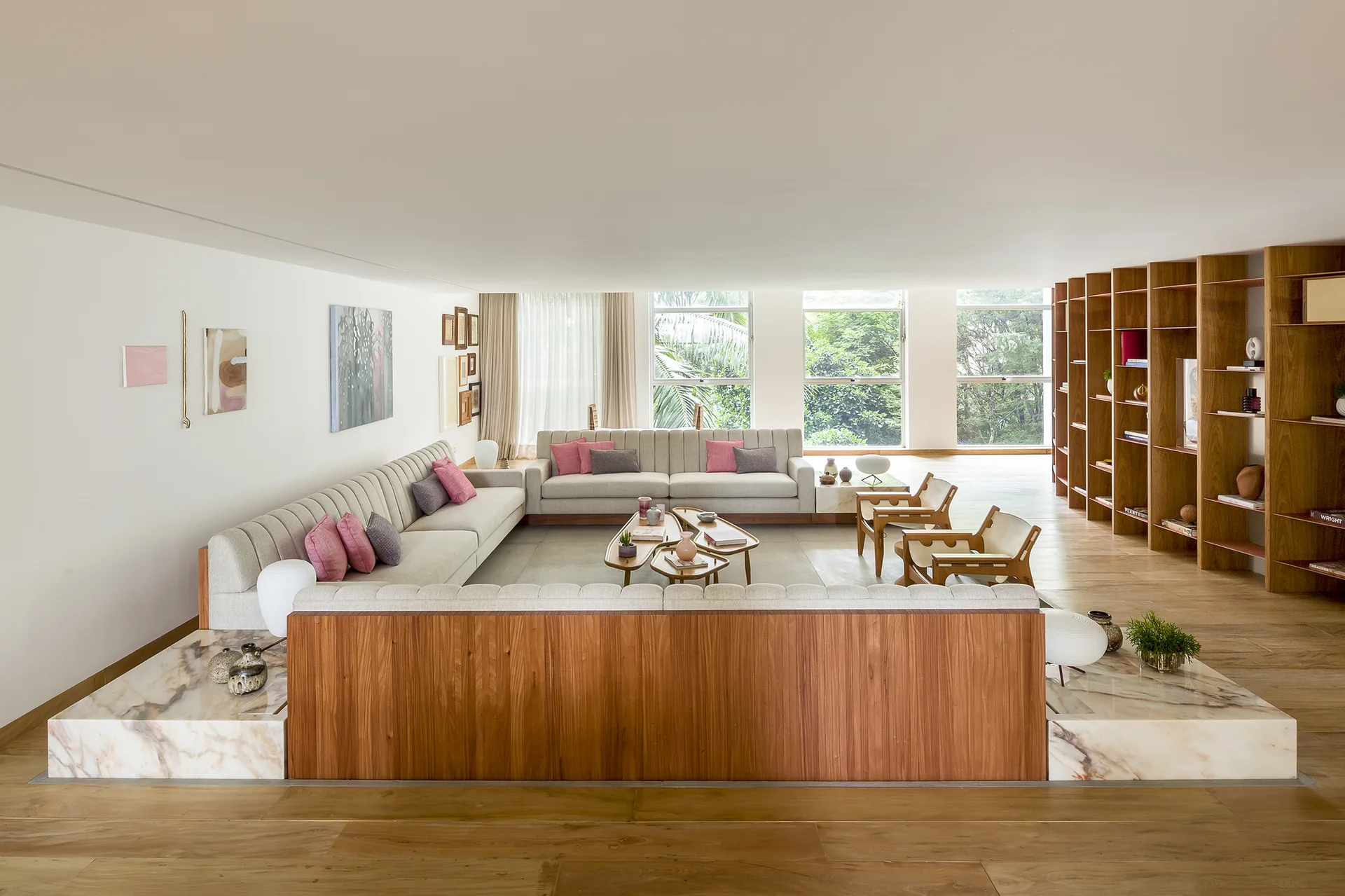 A spacious open-plan living room features a large U-shaped cream sofa set on a veined marble plinth, wooden bookshelves, and wooden coffee tables.