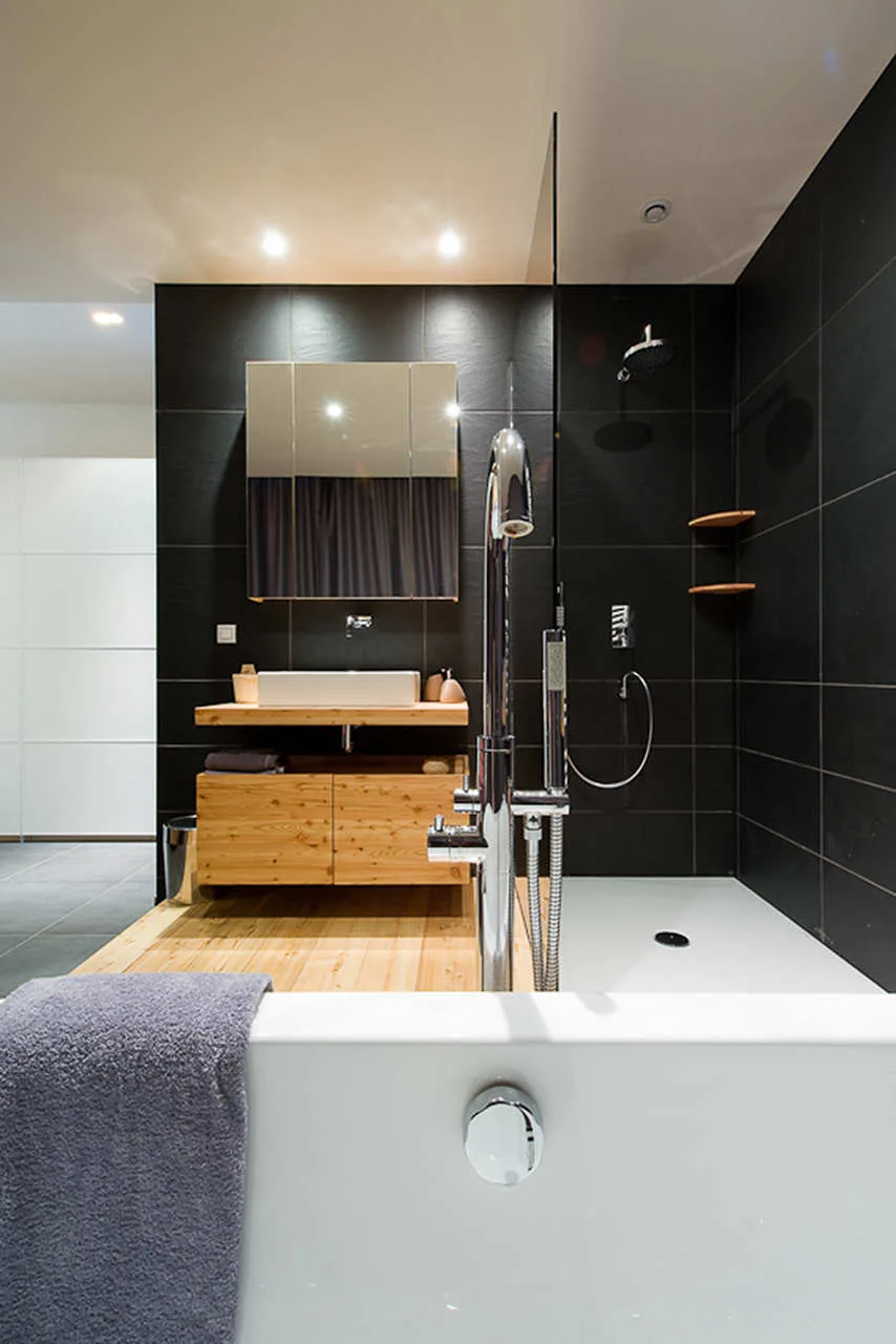 A modern bathroom featuring matte black tiled walls, a light wood floating vanity with a vessel sink, and a white bathtub in the foreground.