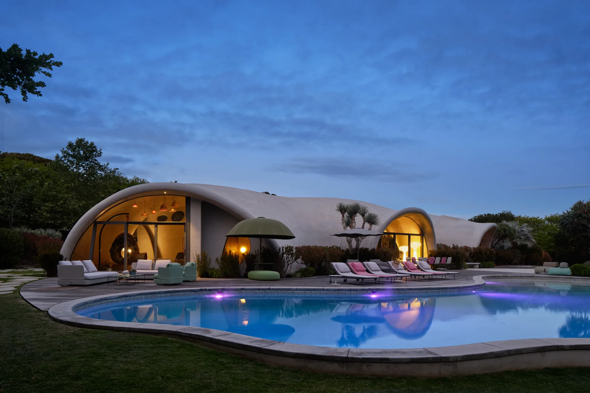 A curved concrete shell house stands next to a lit swimming pool and a wooden deck with lounge chairs at twilight.