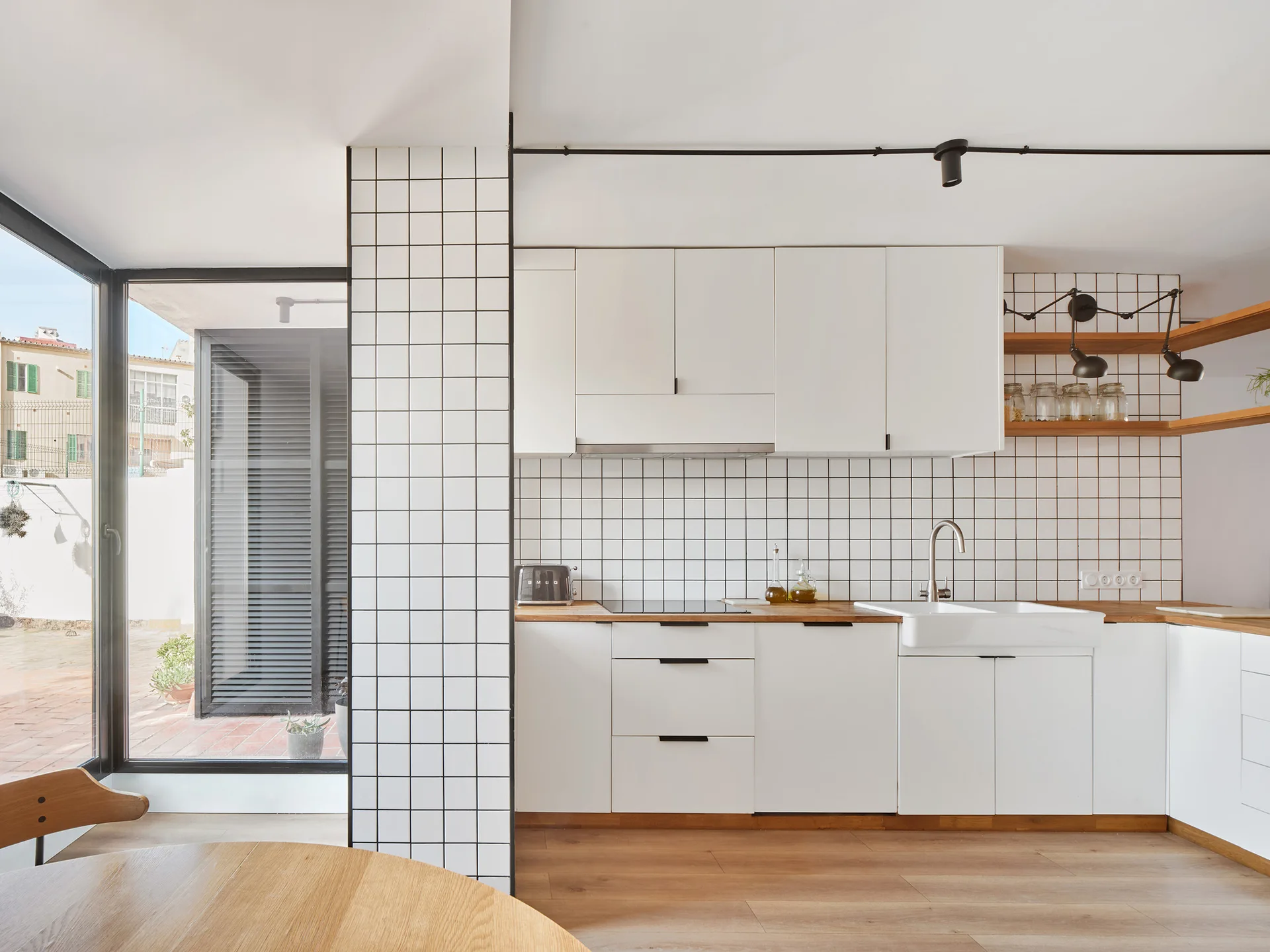 A bright kitchen features white minimalist cabinets, wood countertops, white grid tile walls, and a large glass door leading to an outdoor patio.