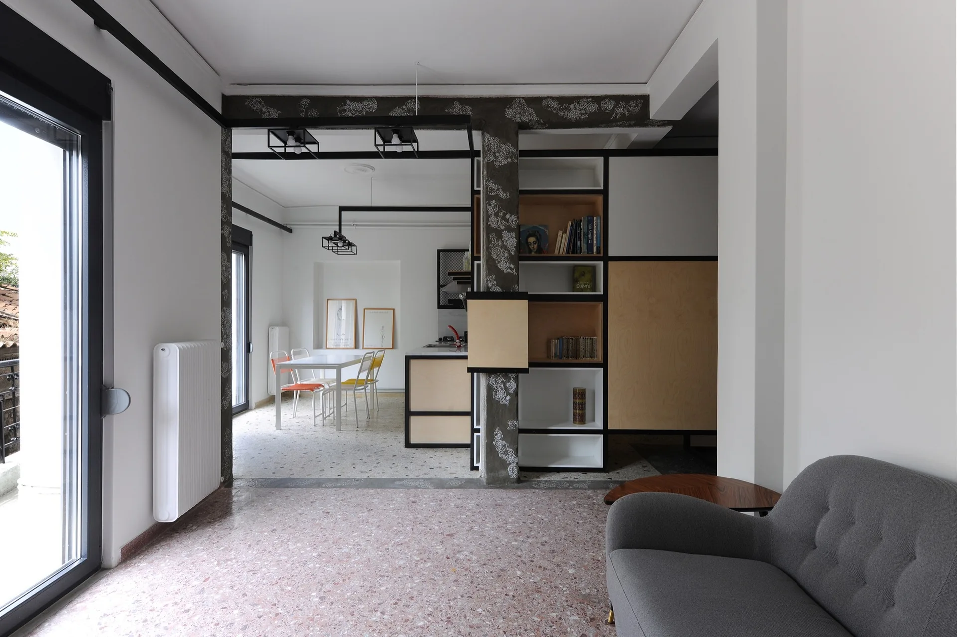 A modern open-plan living room featuring a grey sofa, terrazzo flooring, and a black-framed plywood shelving unit built around a column.