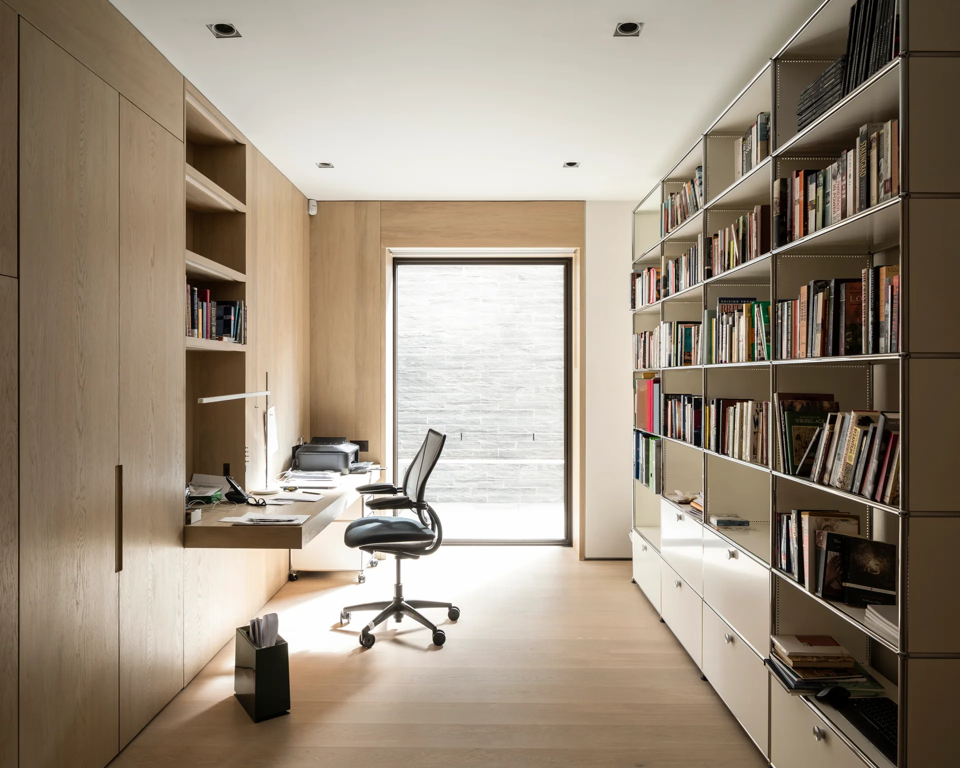 A minimalist home office features light wood built-in cabinets with an integrated desk, a black desk chair, and a modular metal shelving unit filled with books.