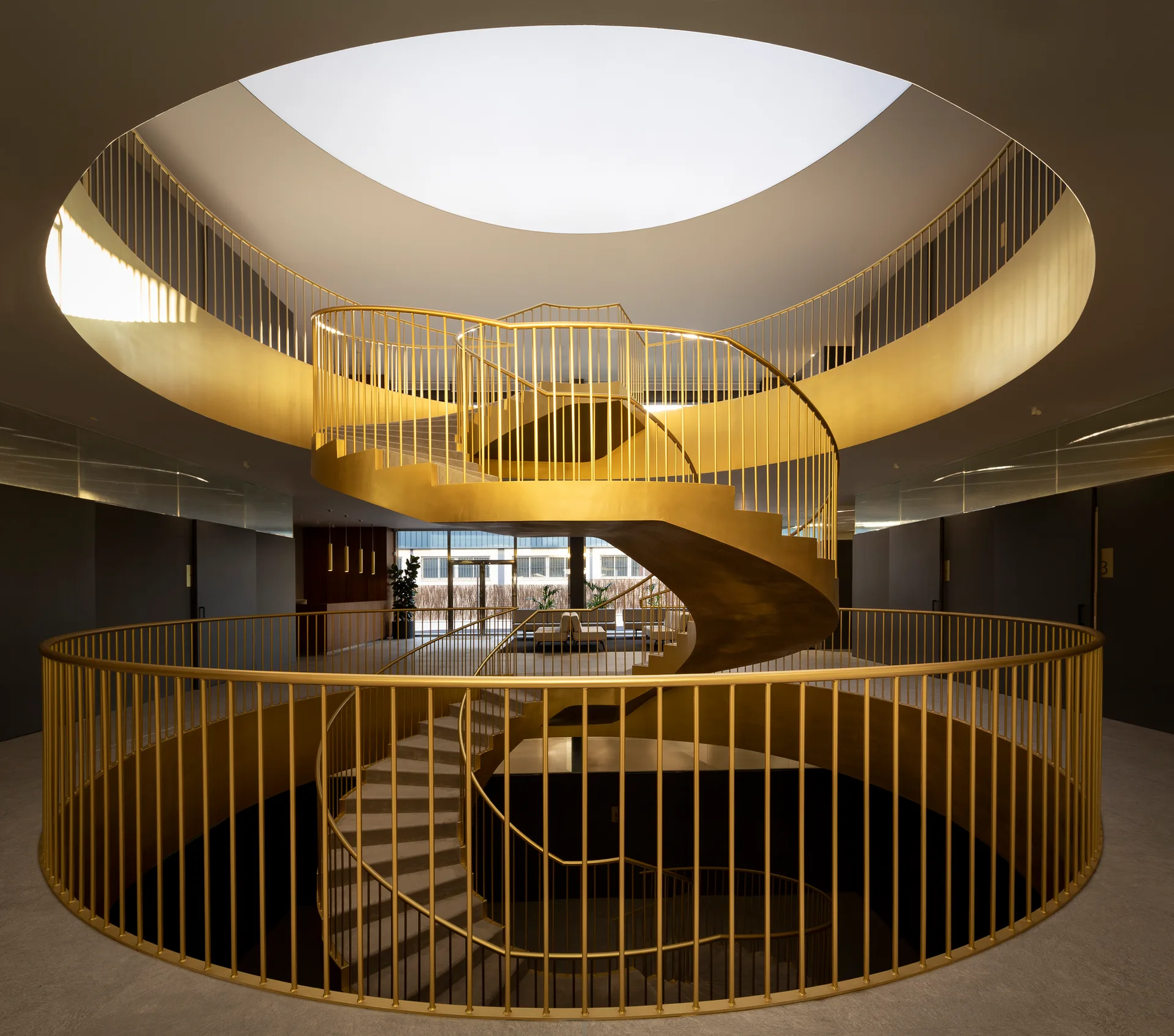 A multi-level gold spiral staircase with vertical metal balusters stands beneath a massive circular skylight in a contemporary gray atrium.