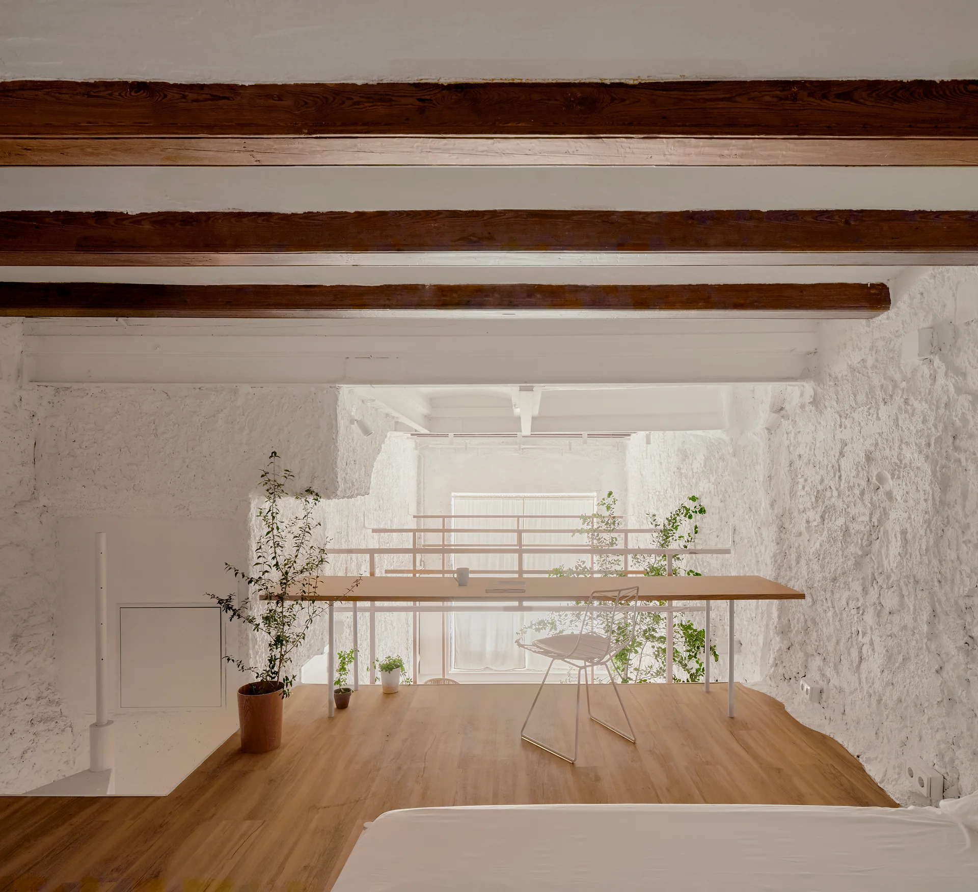 A minimalist loft workspace features a light wood desk, wire chair, and potted plants on wooden floors under exposed dark ceiling beams.