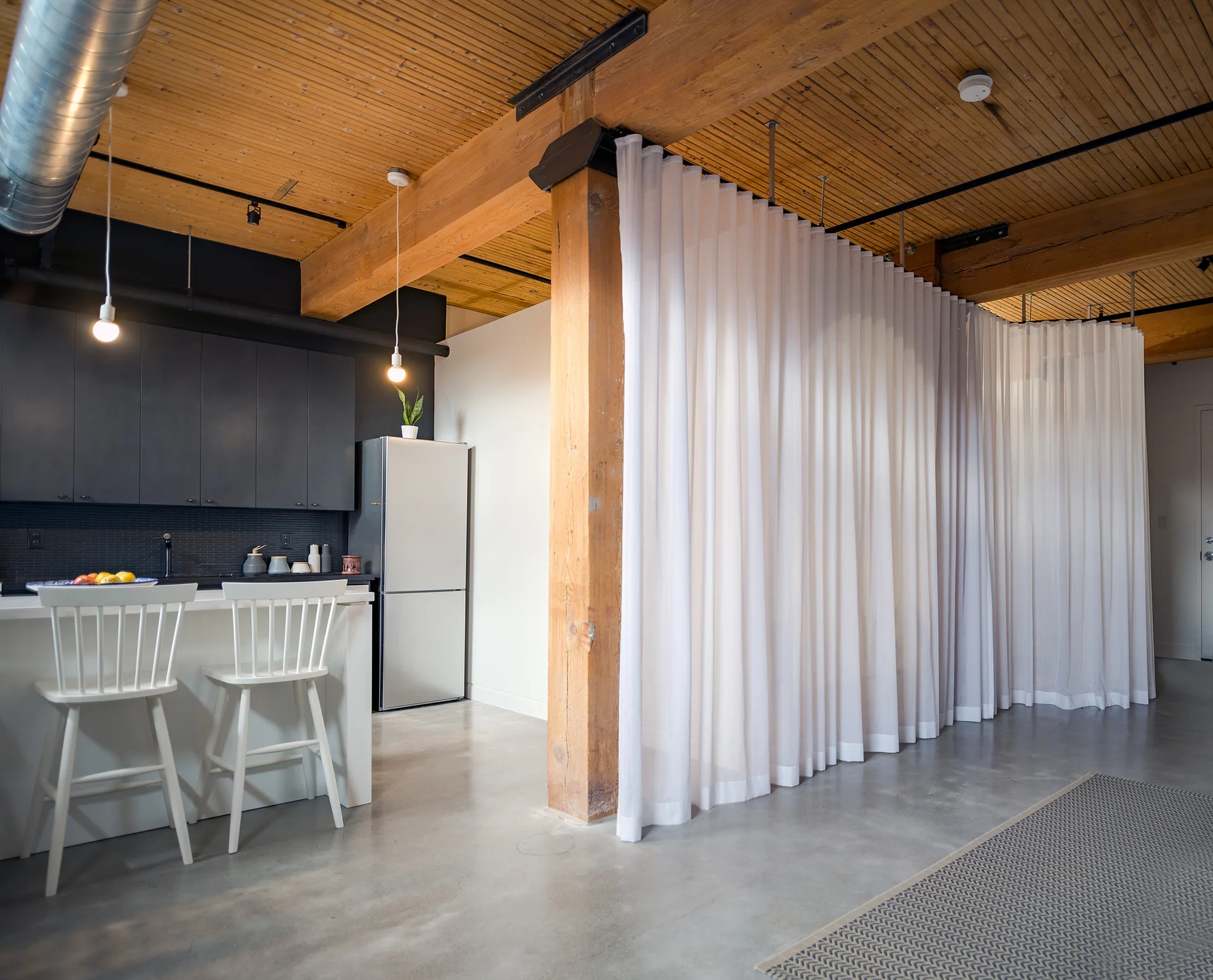 An open-concept loft kitchen features black cabinets, white bar stools, and a tall white curtain divider suspended under heavy timber ceiling beams.