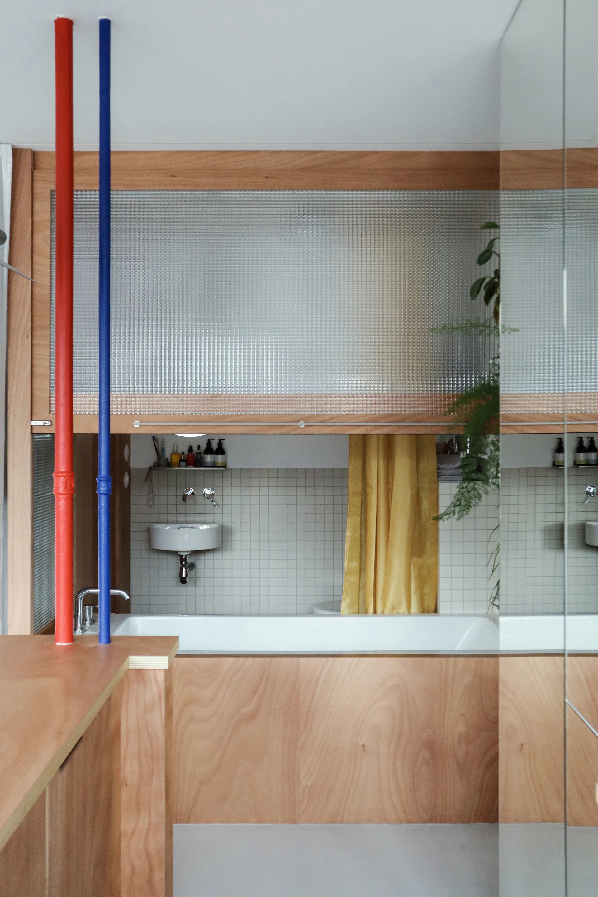A modern bathroom featuring a bathtub encased in wood paneling, white grid-tiled walls, a small floating sink, and vertical red and blue accent poles.