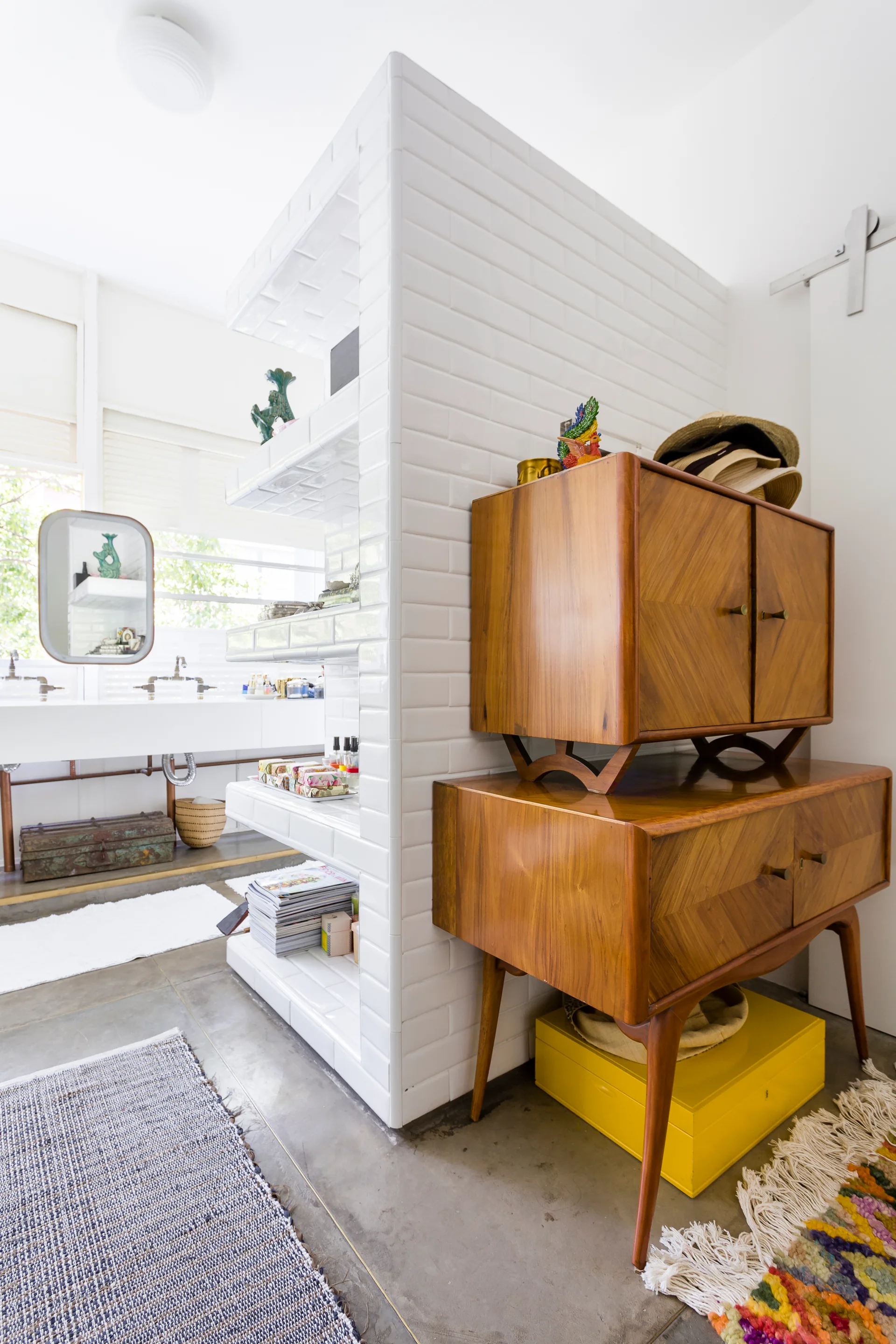 A white subway-tiled partition wall separates a bathroom sink vanity from a mid-century wooden cabinet on a grey concrete floor.