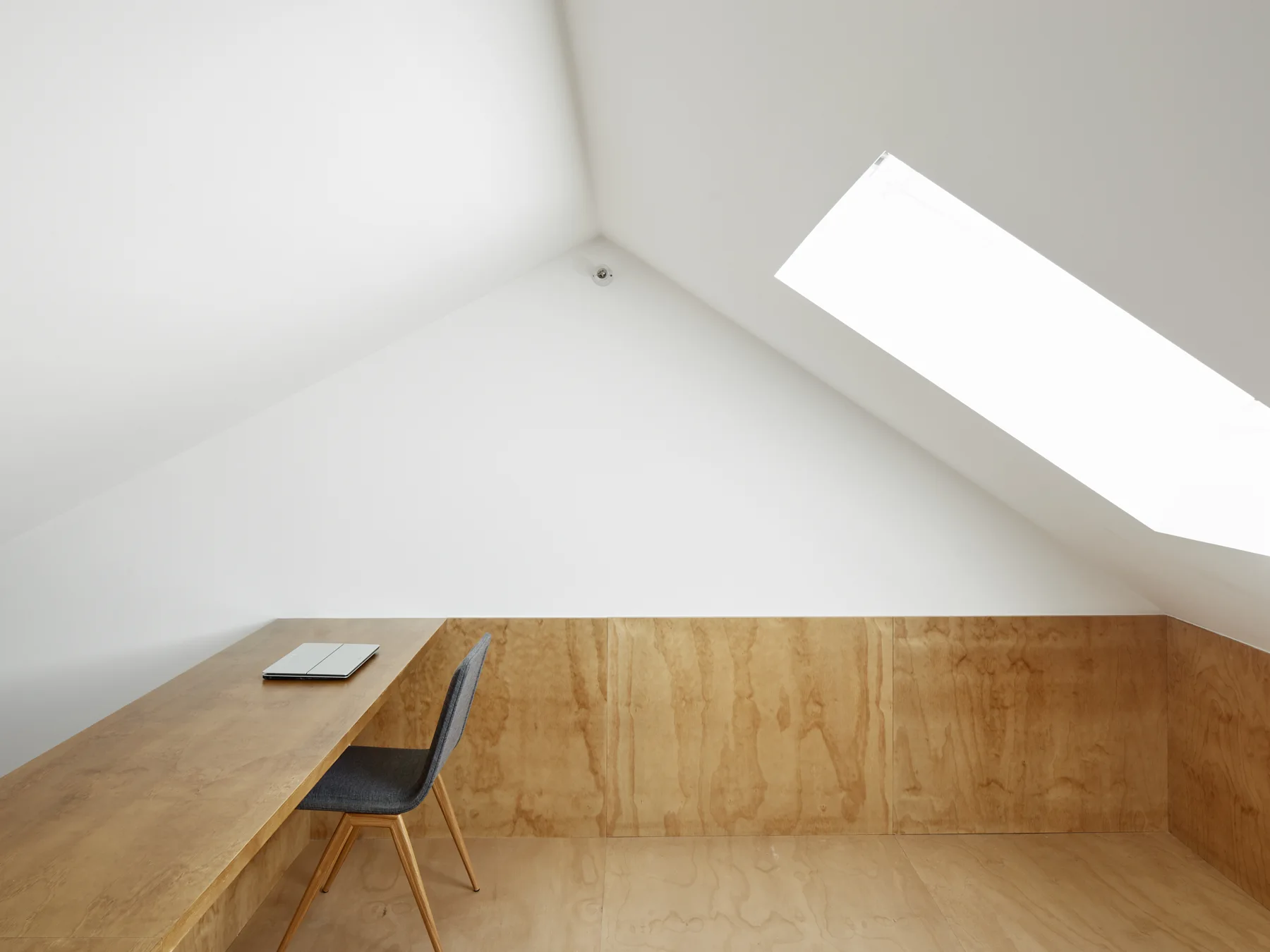 A minimalist attic workspace featuring a long wood desk, a grey chair, and plywood wall panels under a sloped white ceiling with a skylight.