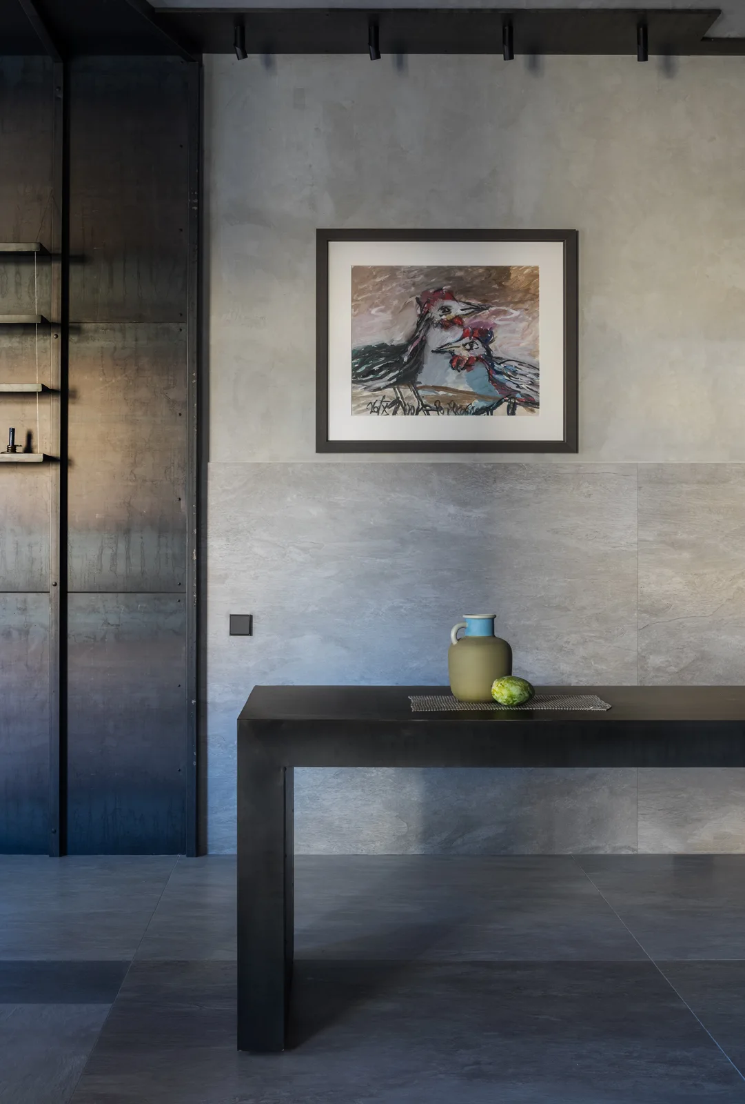 A black metal console table stands against a textured grey stone wall beneath a framed bird painting in an industrial-style space.