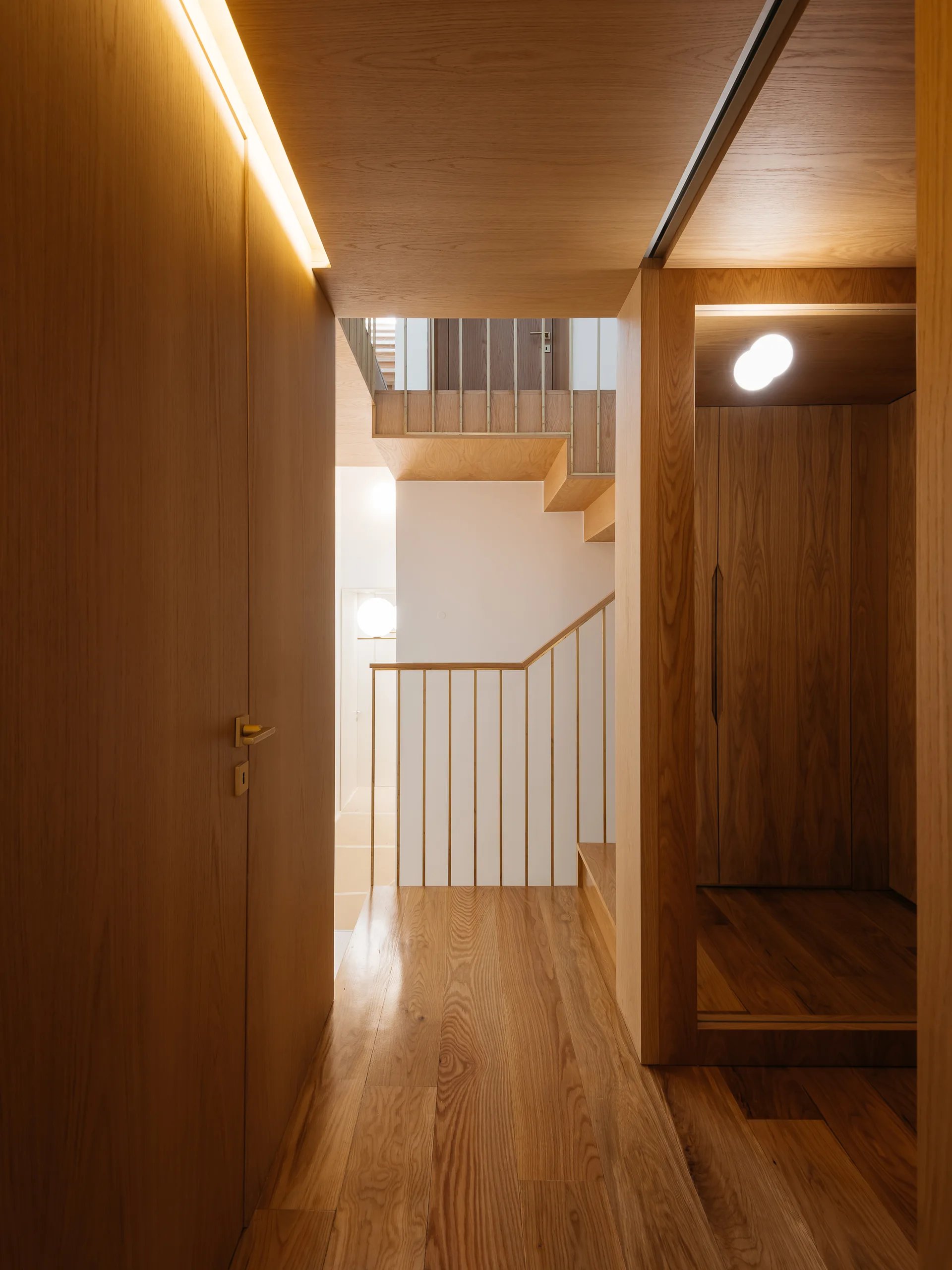 A modern hallway clad entirely in warm wood paneling, featuring a linear cove light, a recessed closet, and a staircase with metal railings.