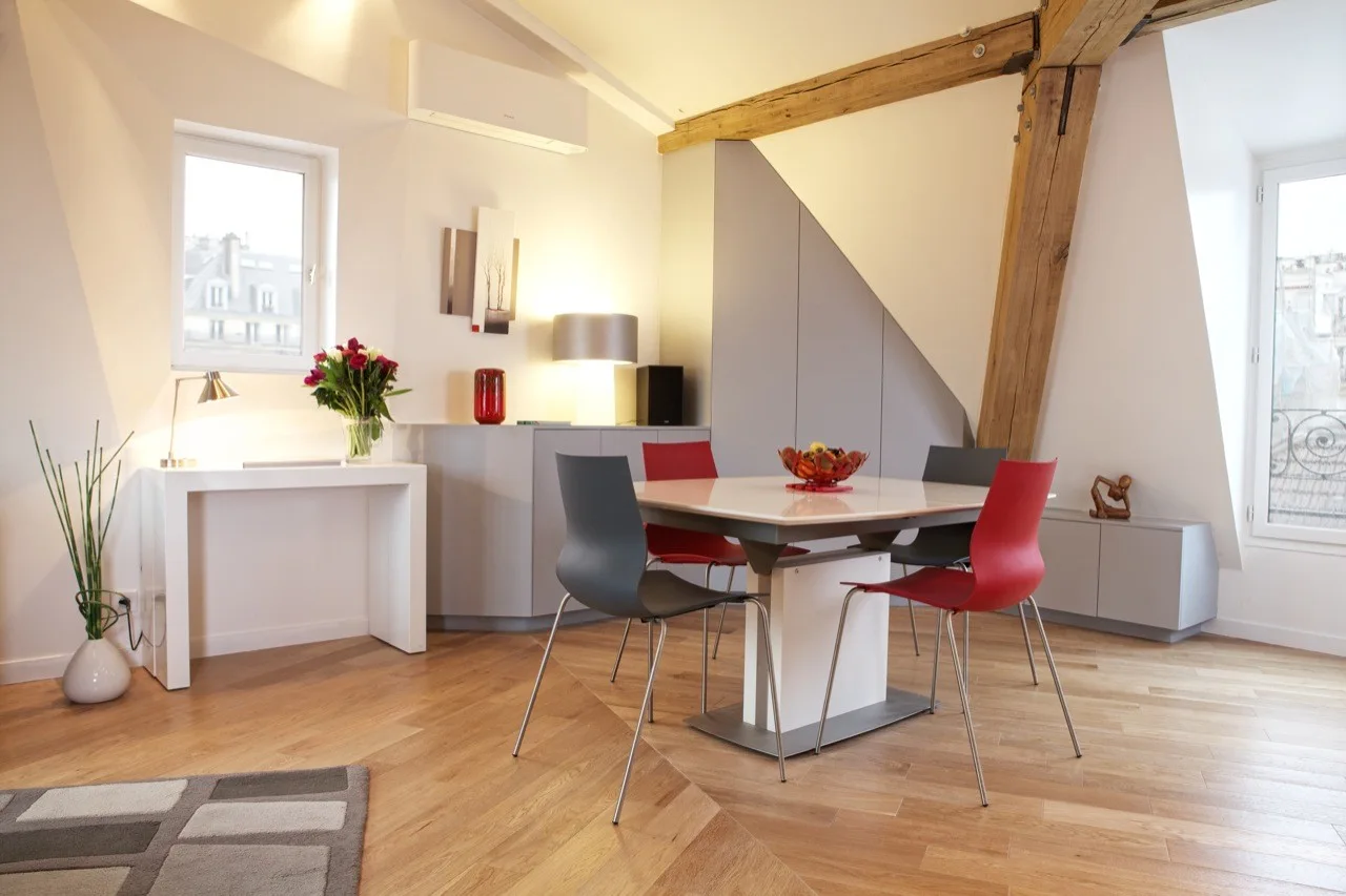 A modern attic dining area features a white square table with red and grey chairs under a sloped ceiling and an exposed wooden beam.